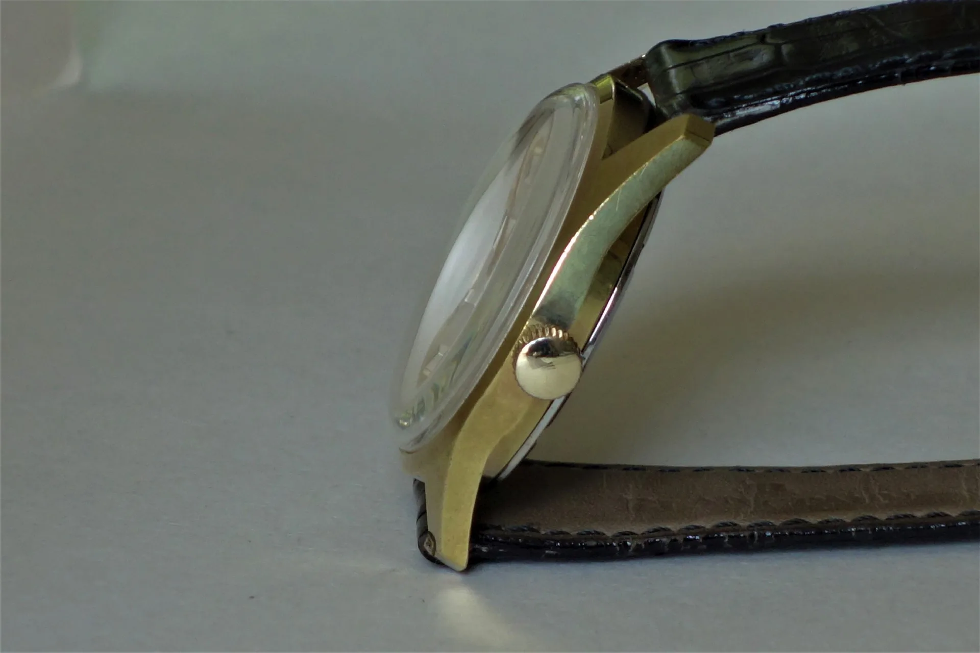Flache Armbanduhr mit goldfarbenem Gehäuse und schwarzem Lederarmband in Seitenansicht.