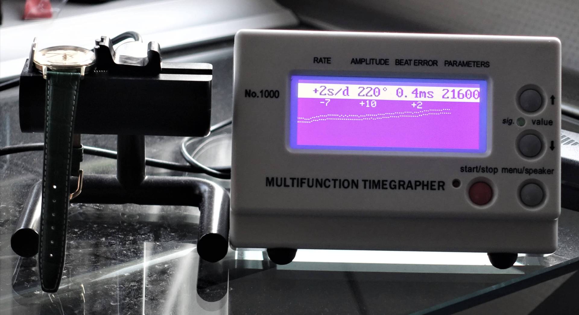 Digitales Uhrentestgerät mit angeschlossener Armbanduhr zur Messung von Ganggenauigkeit und Amplitude.