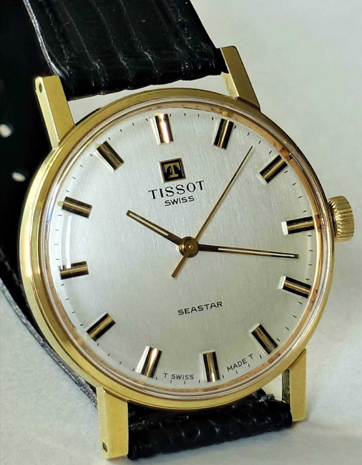 Goldfarbene Armbanduhr mit silberfarbenem Zifferblatt und schwarzem Armband.