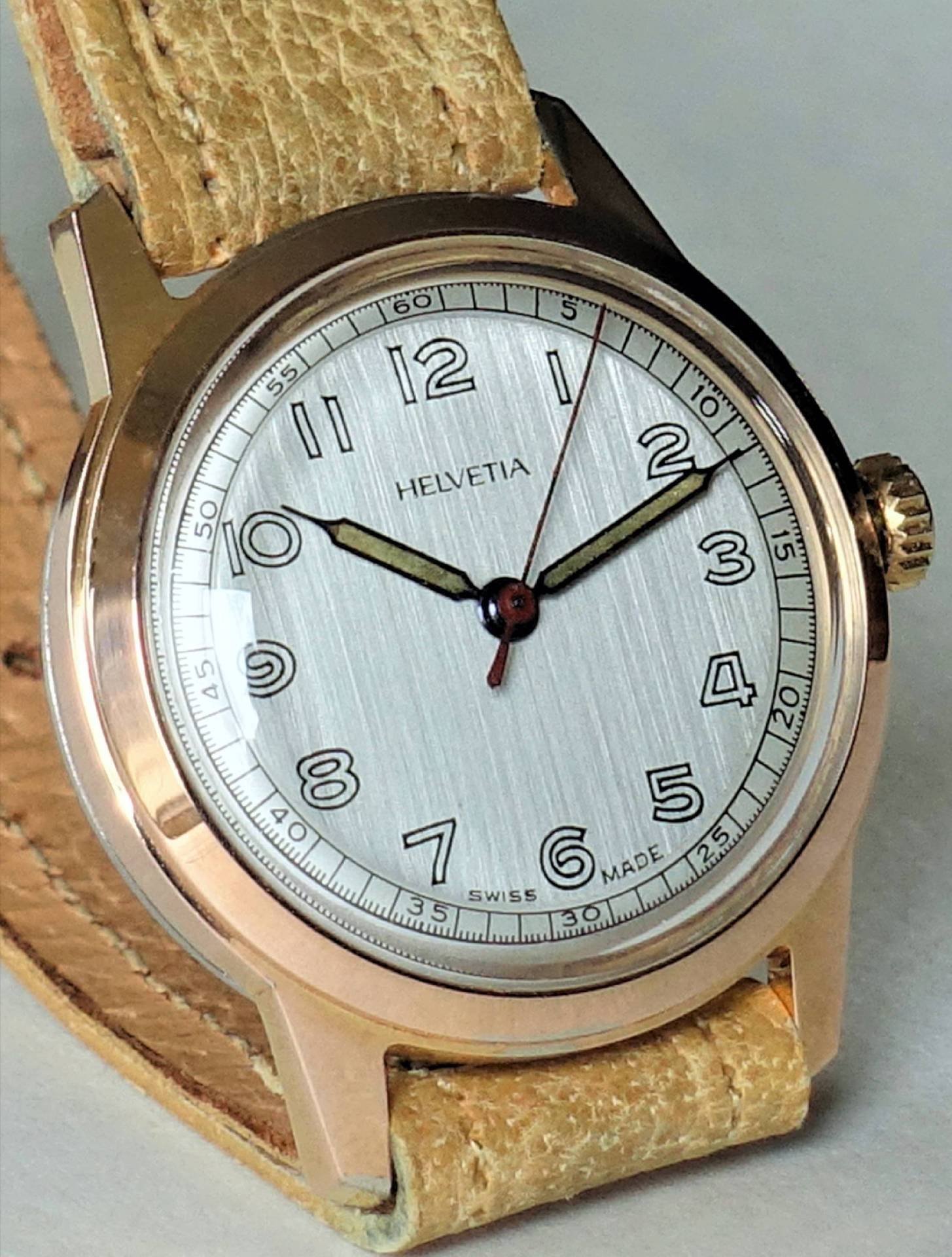 Beige Armbanduhr mit silberfarbenem Zifferblatt und goldfarbenem Gehäuse, Markenname Helvetia auf dem Zifferblatt.