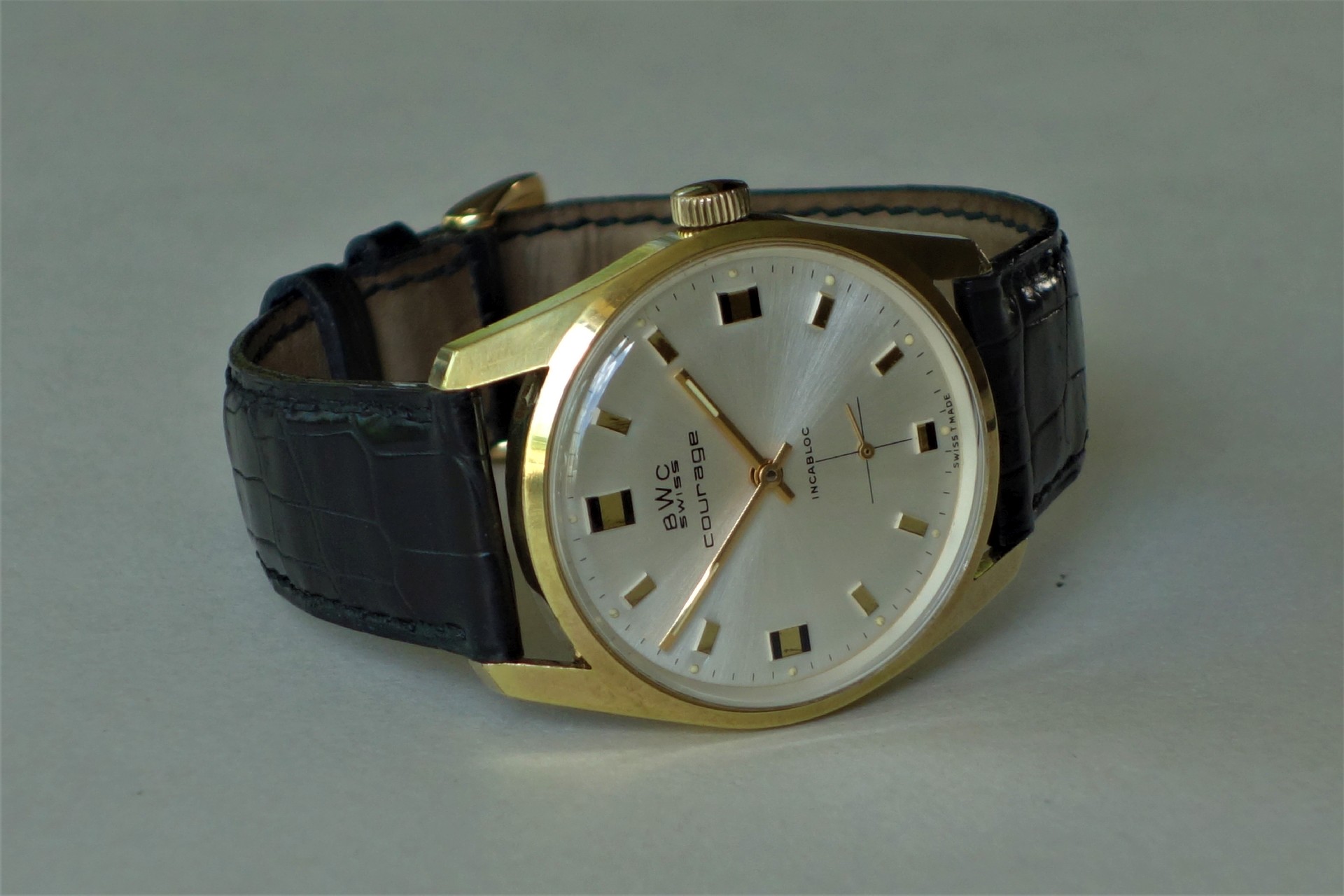 Analoge Armbanduhr mit goldfarbenem Gehäuse, silbernem Zifferblatt und schwarzem Lederarmband.