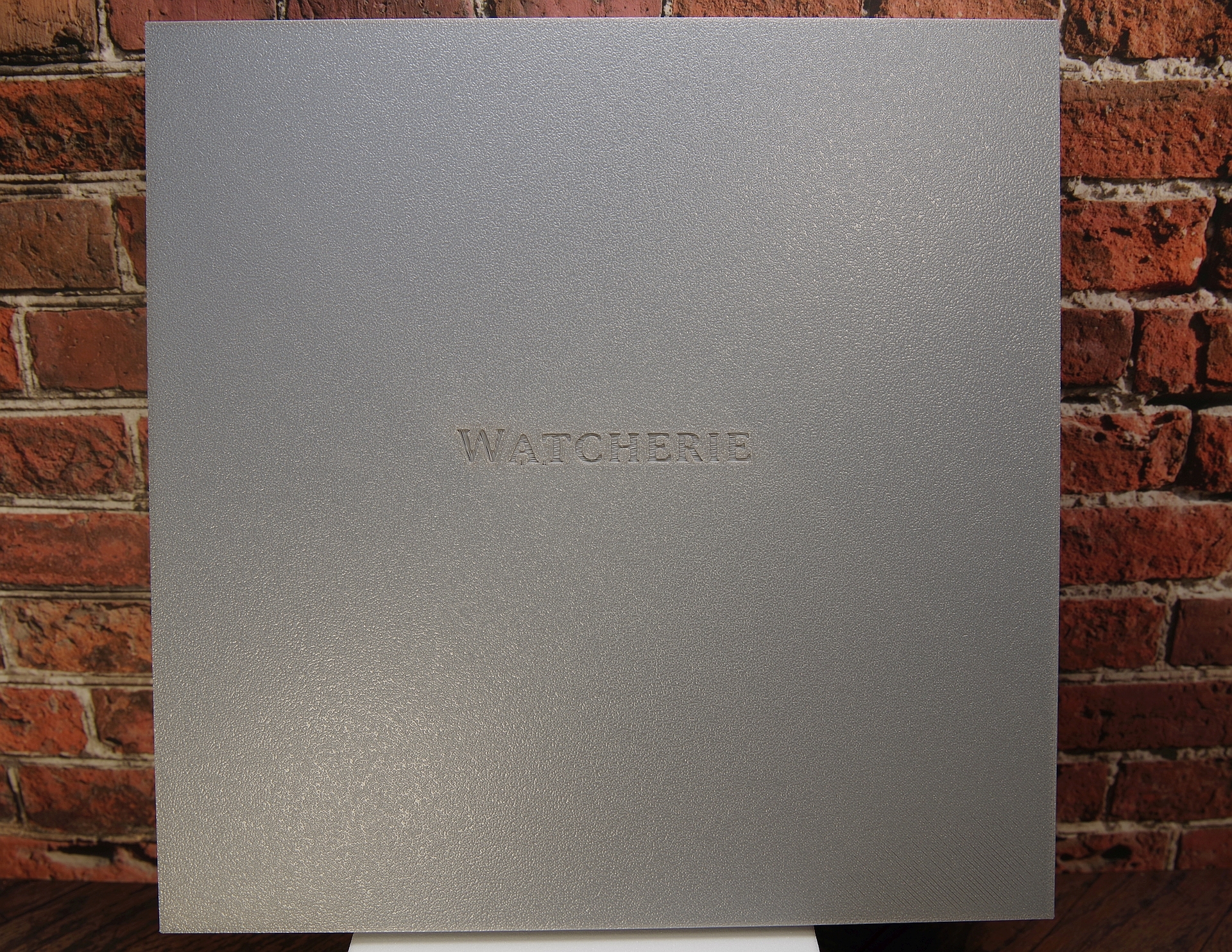 Quadratische silberfarbene Uhrenbox mit geprägtem Schriftzug "WATCHERIE" auf der Vorderseite.