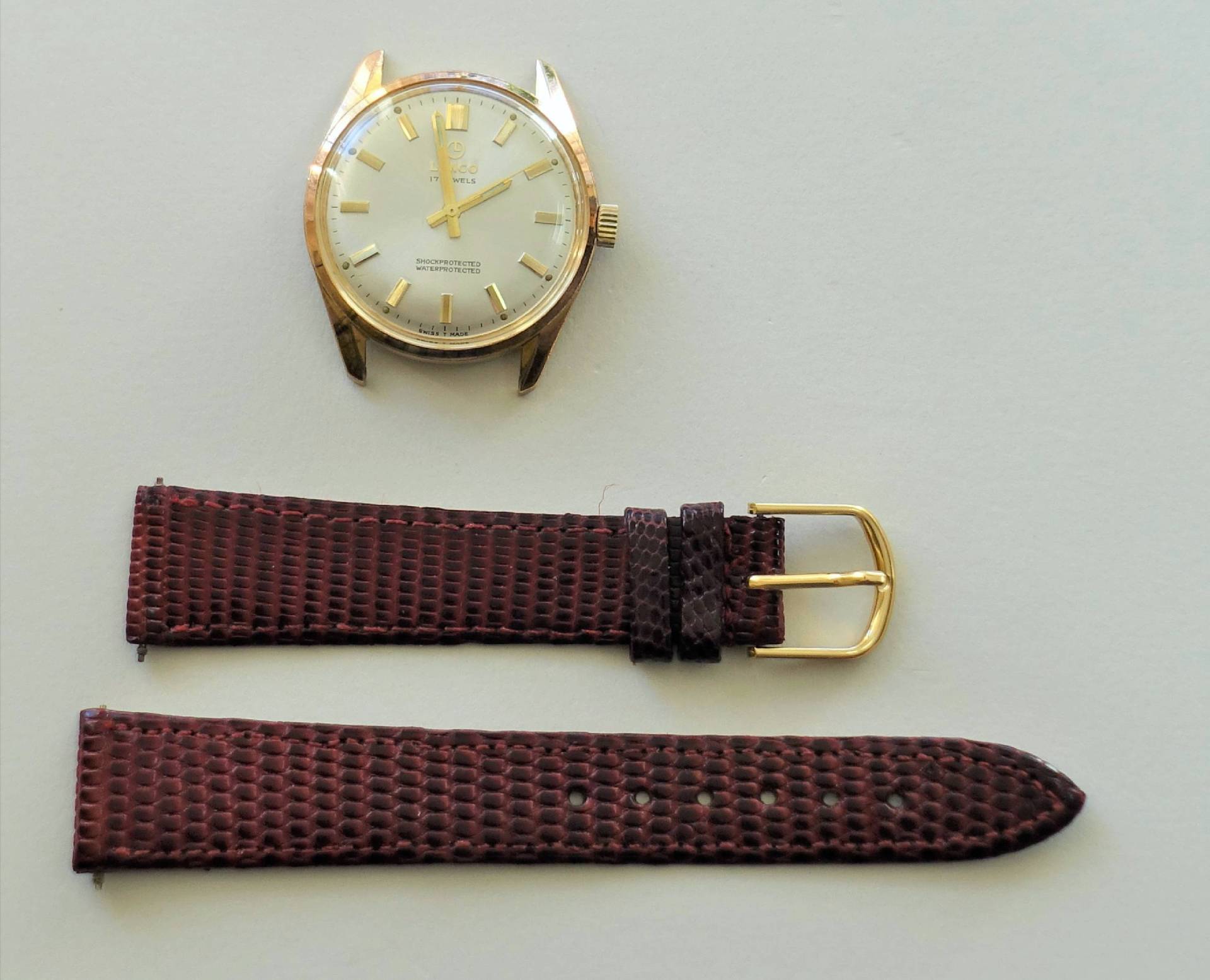 Goldfarbene Armbanduhr mit hellem Zifferblatt und separat liegendem braunen Lederarmband mit Schließe.