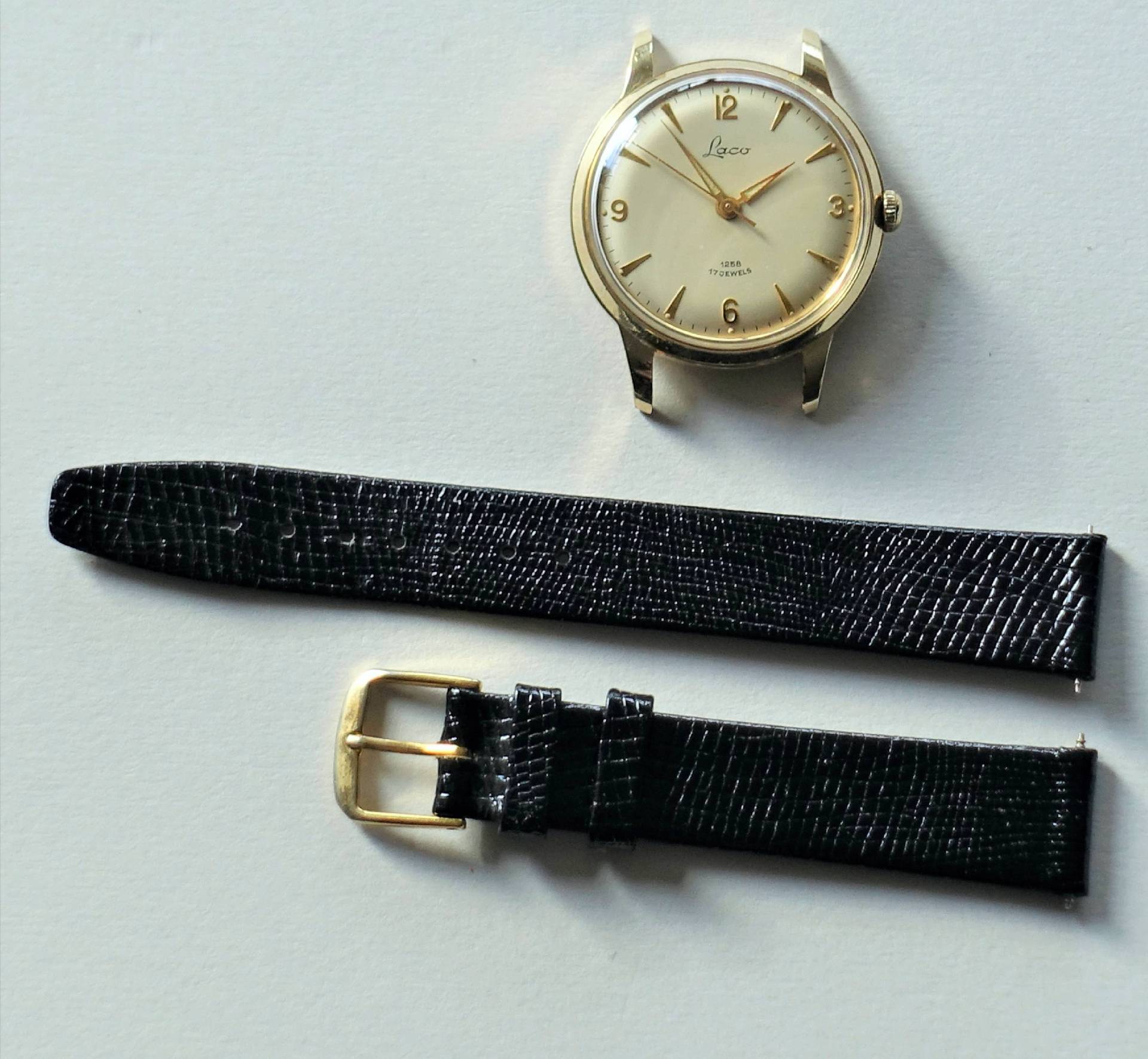 Goldfarbene Armbanduhr mit hellem Zifferblatt und schwarzem Lederarmband in zwei Teilen.