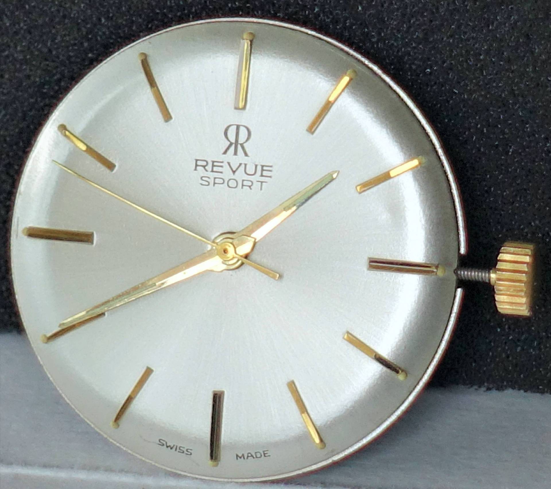 Rundes silberfarbenes Uhrwerk mit goldfarbenen Zeigern und Indizes, sichtbarer Krone.