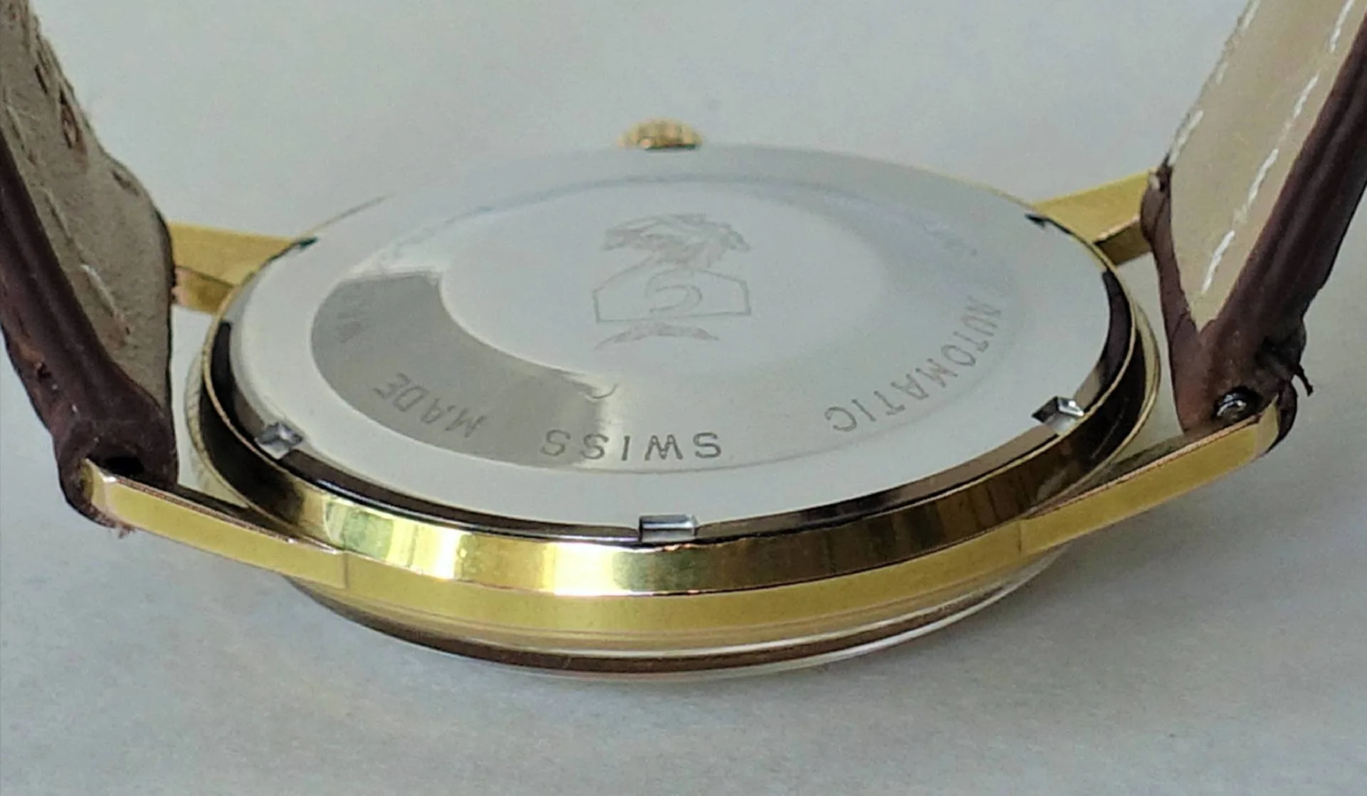 Die Nahaufnahme zeigt die Rückseite einer Armbanduhr mit silberfarbenem Gehäuseboden, goldfarbenem Gehäuserand und braunem Lederarmband.