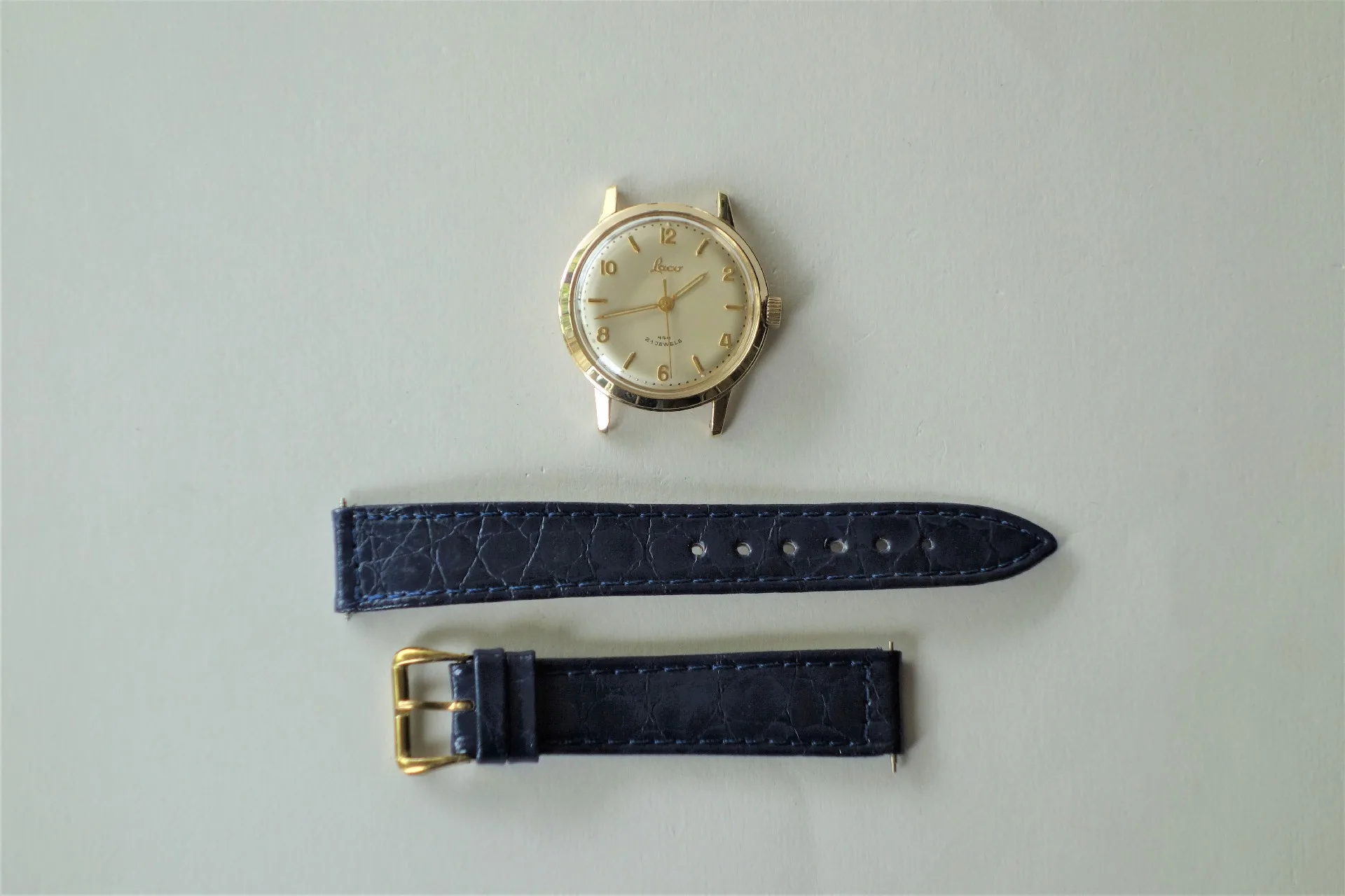 Uhrgehäuse goldfarben Rundes Uhrgehäuse in goldfarbener Optik mit hellem Zifferblatt und goldfarbenen Zeigern. Titel: Uhrenarmband dunkelblau Alt-Text: Zweiteiliges Uhrenarmband in dunkelblauer Farbe mit strukturierter Oberfläche und goldfarbener Schließe.