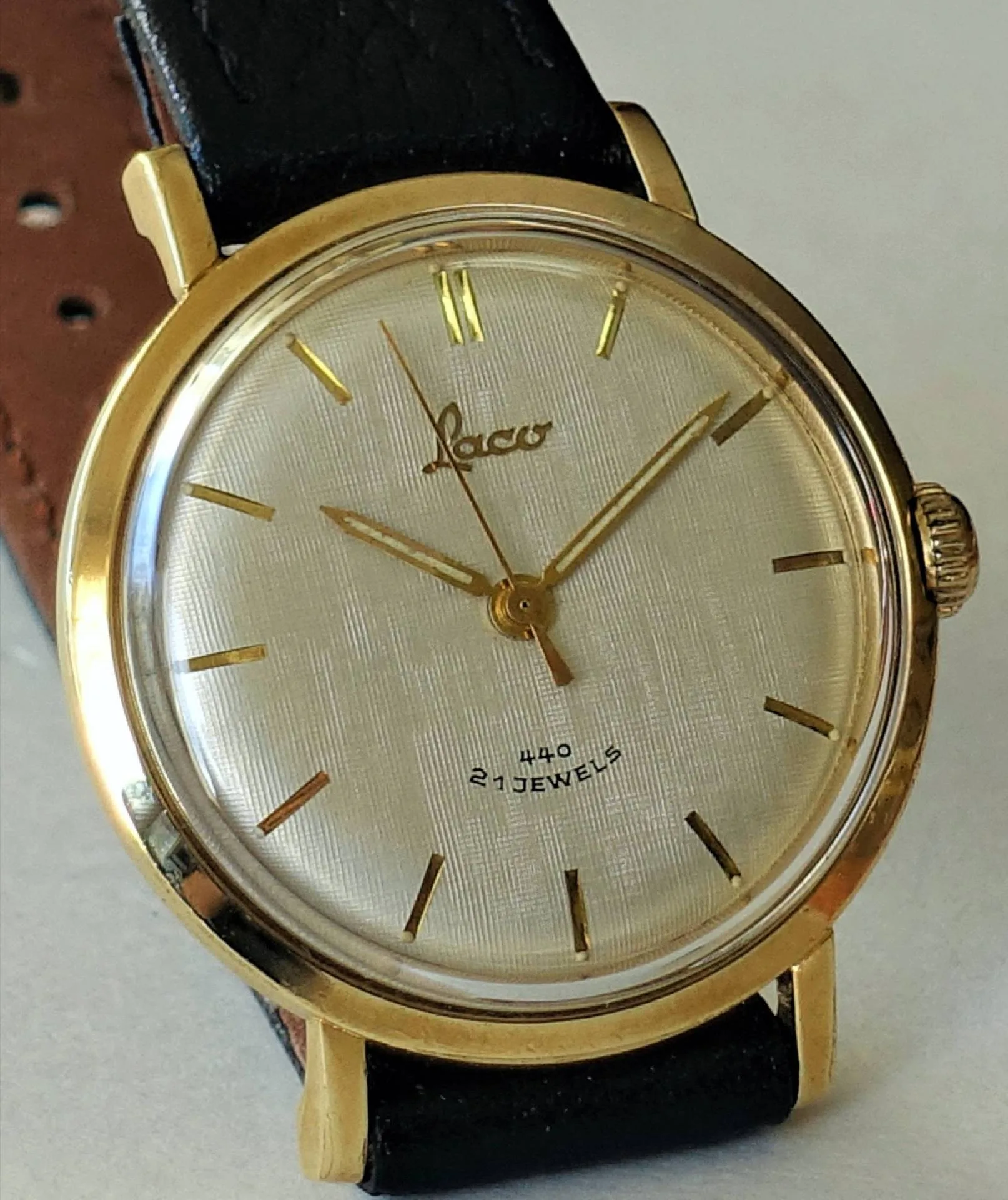 Goldfarbene Armbanduhr mit hellem Zifferblatt, schwarzen Lederarmband und goldenen Zeigern.