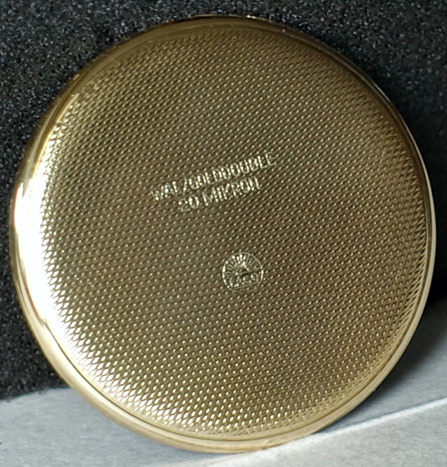 Goldfarbener Uhrendeckel fein strukturiert Rückseite eines goldfarbenen, fein strukturierten Uhrendeckels mit eingeprägter Beschriftung.