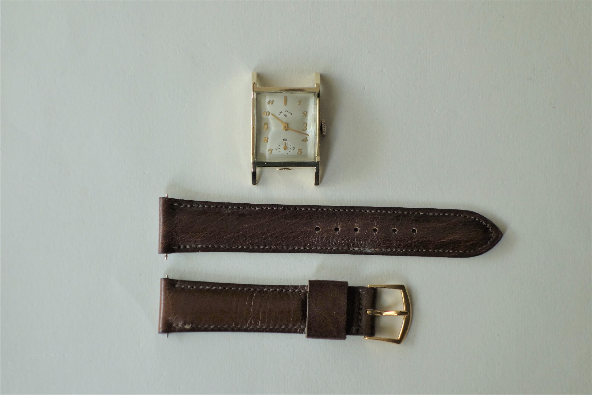Rechteckiges goldfarbenes Uhrgehäuse mit weißem Zifferblatt und goldenen Zahlen, daneben ein braunes Lederarmband in zwei Teilen.