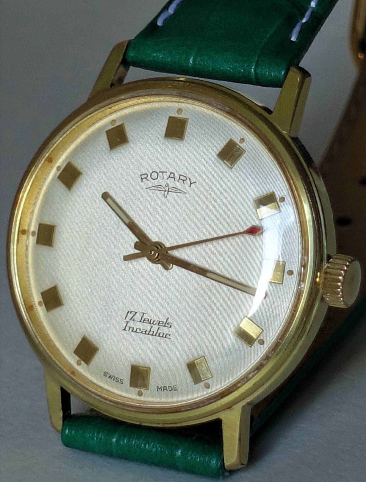 Analoge Armbanduhr mit goldfarbenem Gehäuse, weißem Zifferblatt und grünem Armband.