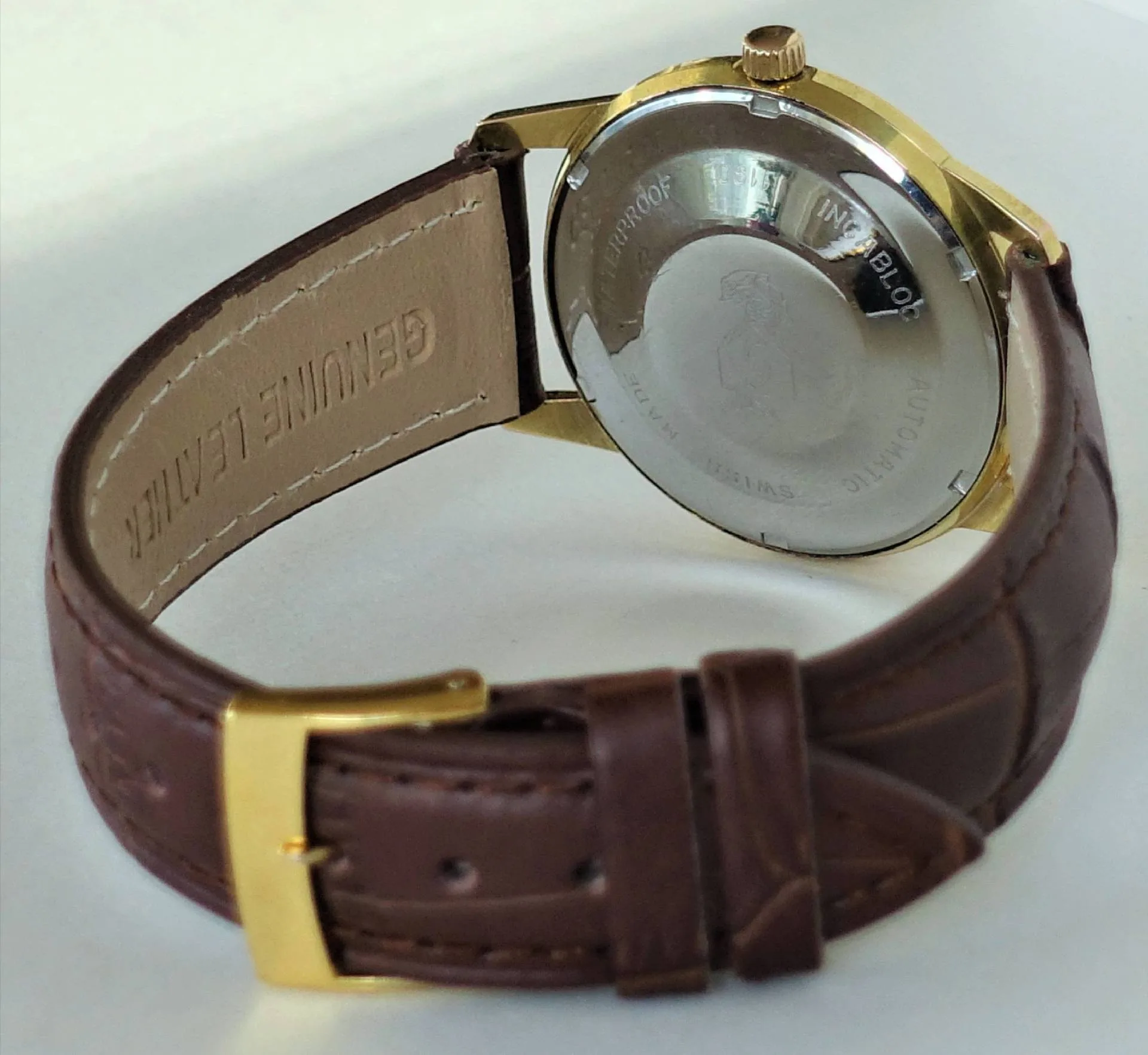 Braun-goldene Uhr Rückansicht Die Rückseite einer Armbanduhr mit braunem Lederarmband und goldfarbenem Gehäuse ist zu sehen.
