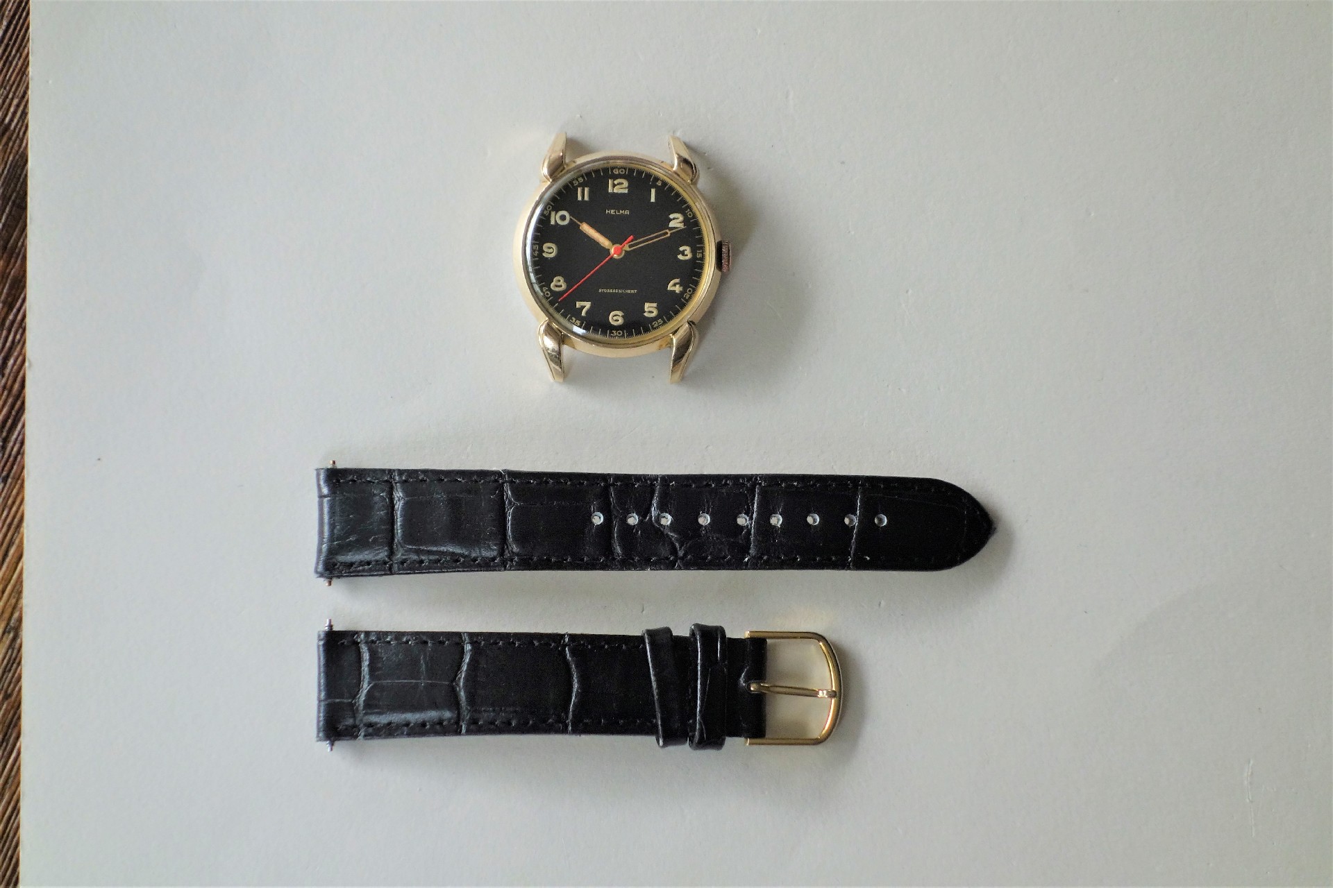 Zweiteiliges schwarzes Uhrenarmband mit goldfarbener Schließe, neben einem goldfarbenen Uhrengehäuse mit schwarzem Zifferblatt.