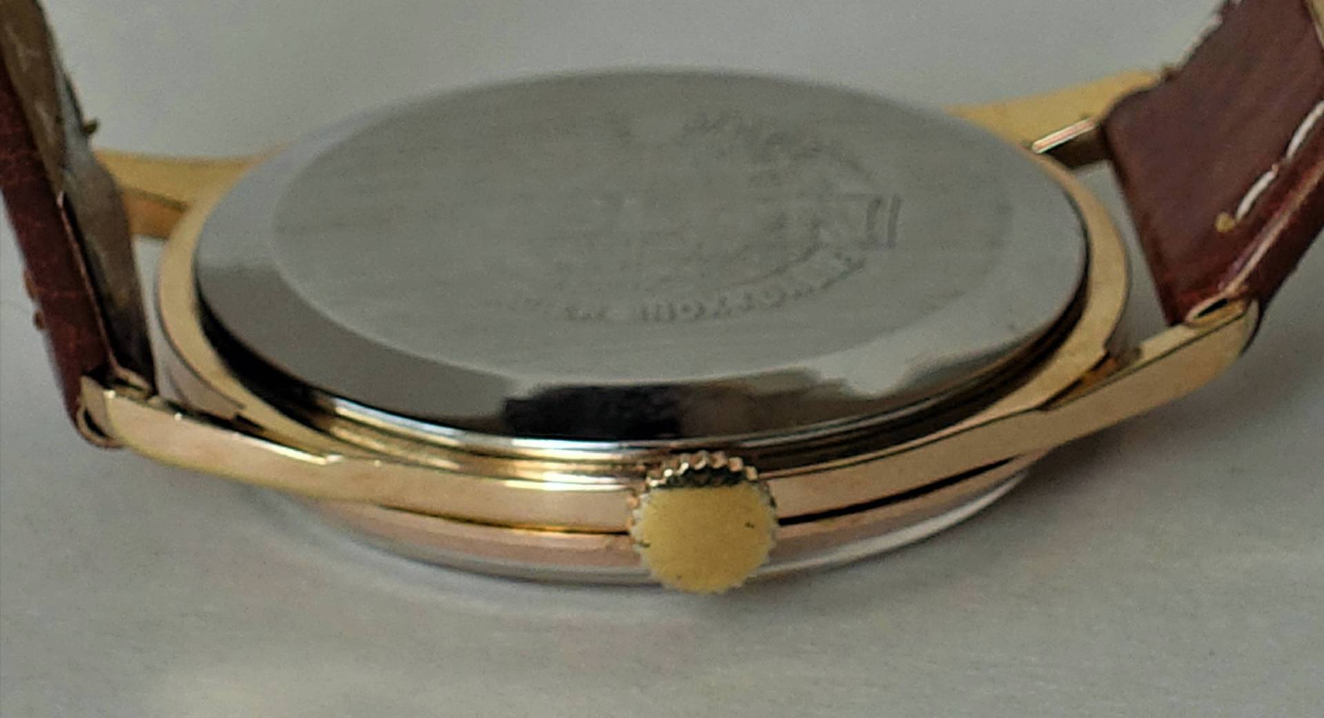 Die Aufnahme zeigt die Rückseite einer Armbanduhr mit silberfarbenem Gehäuseboden, goldfarbenem Gehäuserand und Krone sowie braunem Lederarmband.