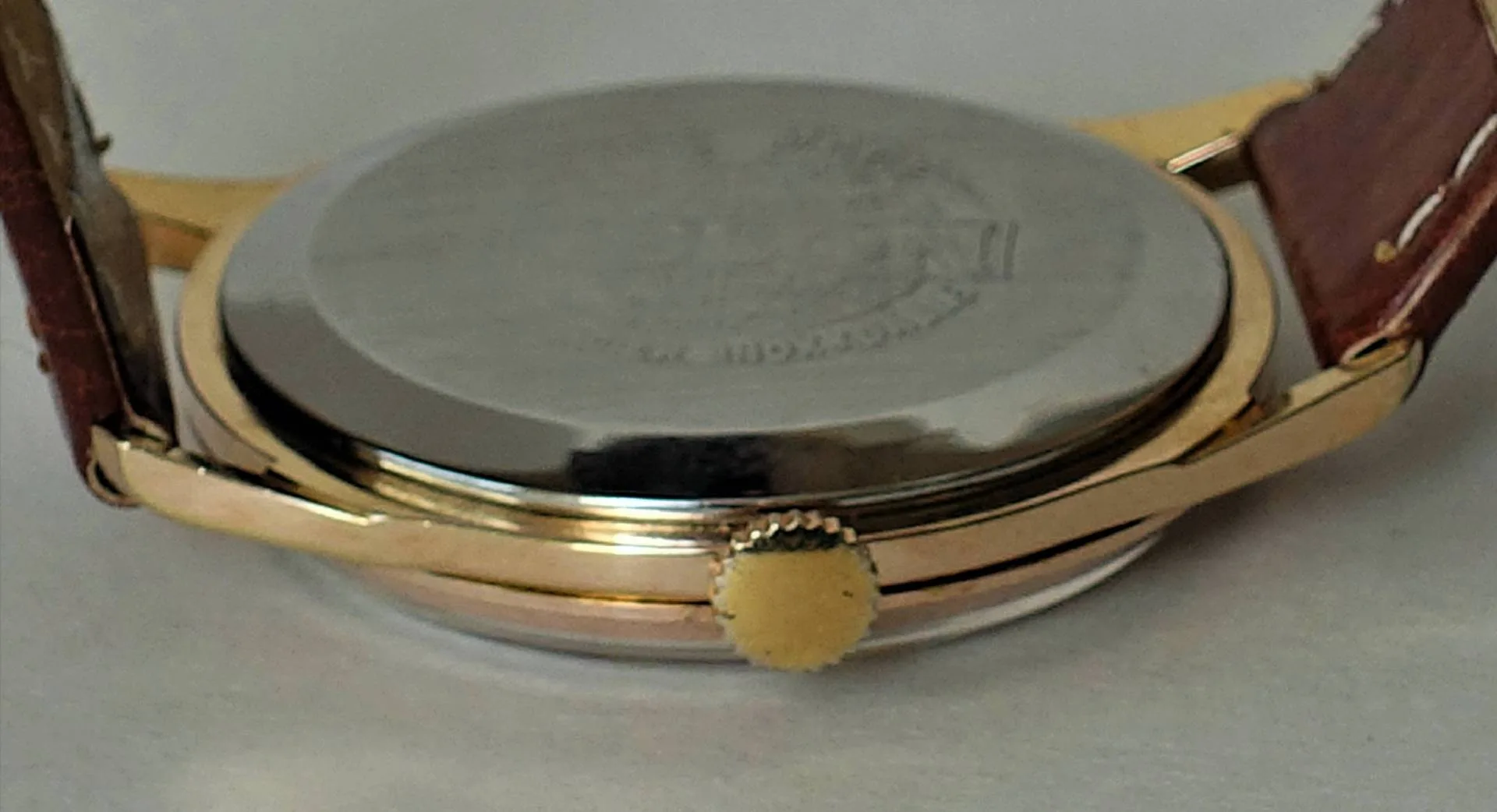 Die Aufnahme zeigt die Rückseite einer Armbanduhr mit silberfarbenem Gehäuseboden, goldfarbenem Gehäuserand und Krone sowie braunem Lederarmband.