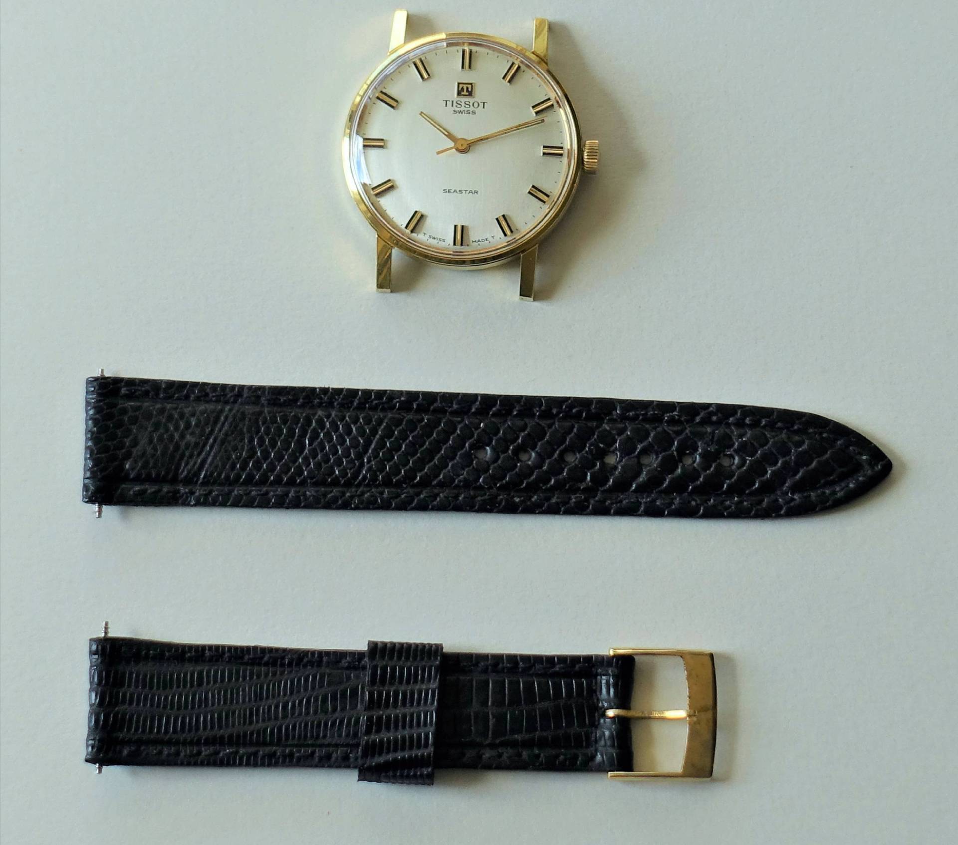Zerlegte Armbanduhr mit rundem goldfarbenem Gehäuse und schwarzem Armband in zwei Teilen.