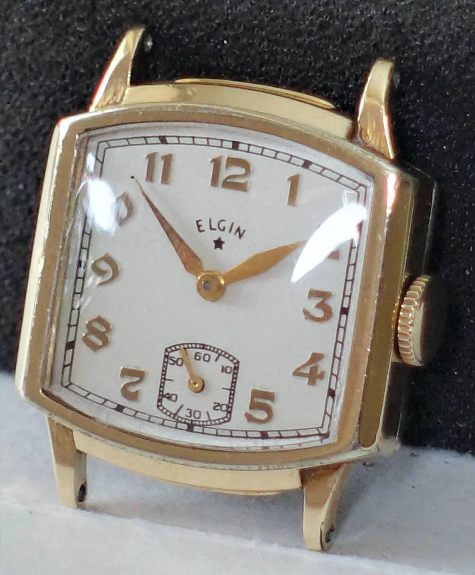 Eine rechteckige Armbanduhr mit goldfarbenem Gehäuse, weißen Zifferblatt und goldfarbenen Zahlen sowie Zeigern, darunter eine kleine Sekundenanzeige.