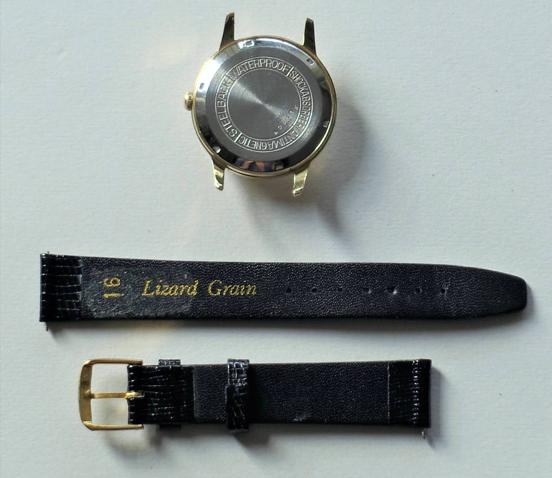 Goldfarbenes Uhrgehäuse mit silberner Rückseite und schwarzes zweiteiliges Uhrenarmband mit goldfarbener Schließe.