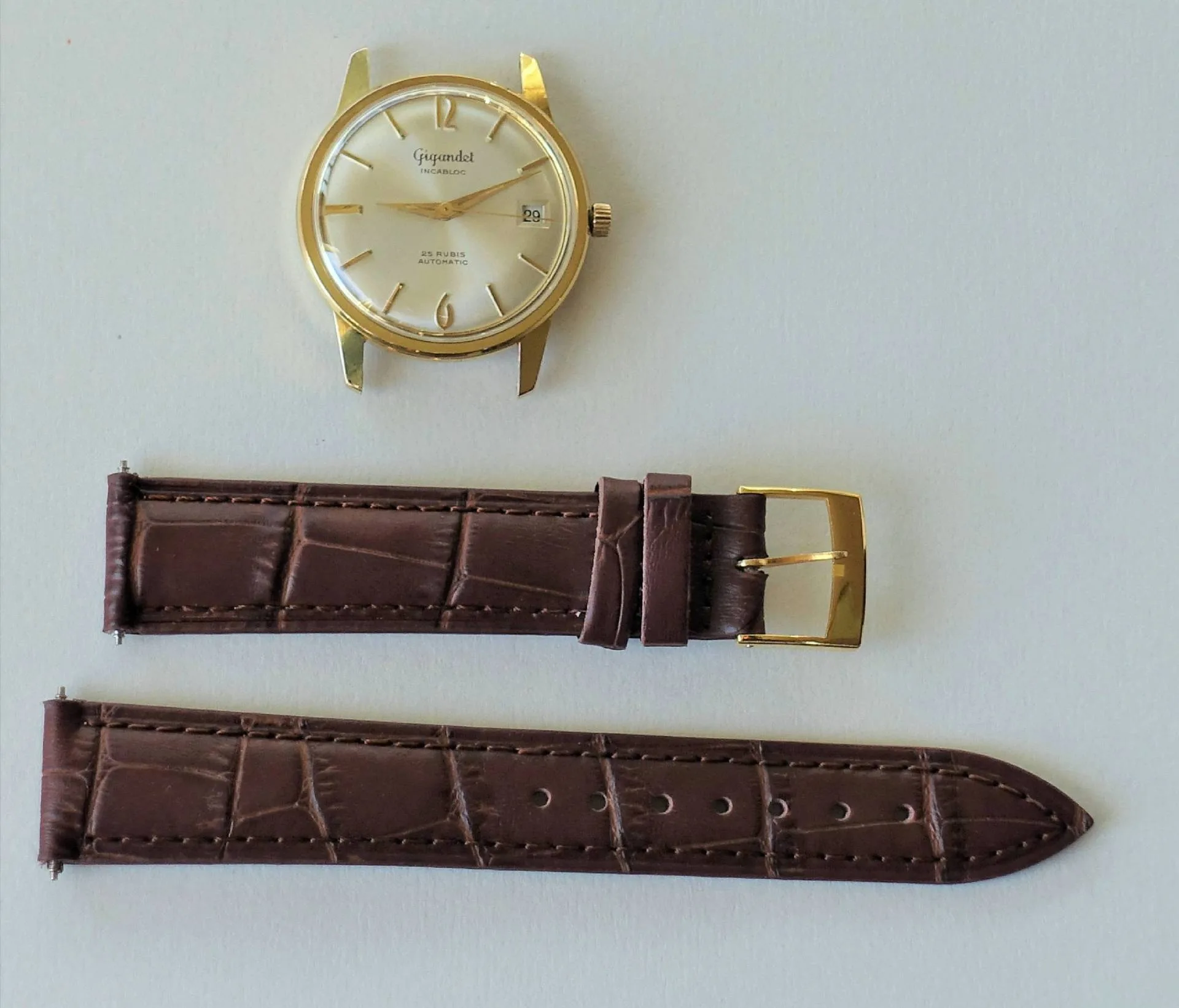 Goldfarbene Uhr mit braunem Lederband Eine goldfarbene Armbanduhr mit hellem Zifferblatt liegt neben einem zweiteiligen braunen Lederarmband mit goldfarbener Schließe.
