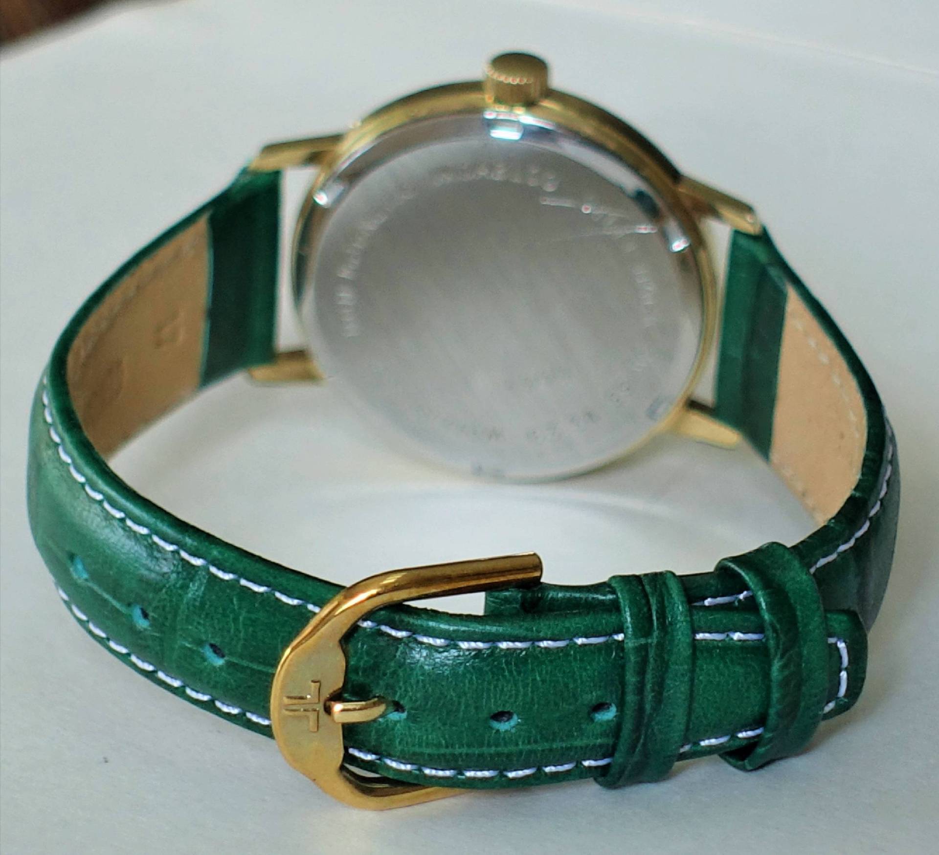 Eine Armbanduhr mit goldfarbenem Gehäuse und grünem Lederarmband, fotografiert von hinten, sodass der verschlossene Gehäuseboden und die goldene Dornschließe sichtbar sind.