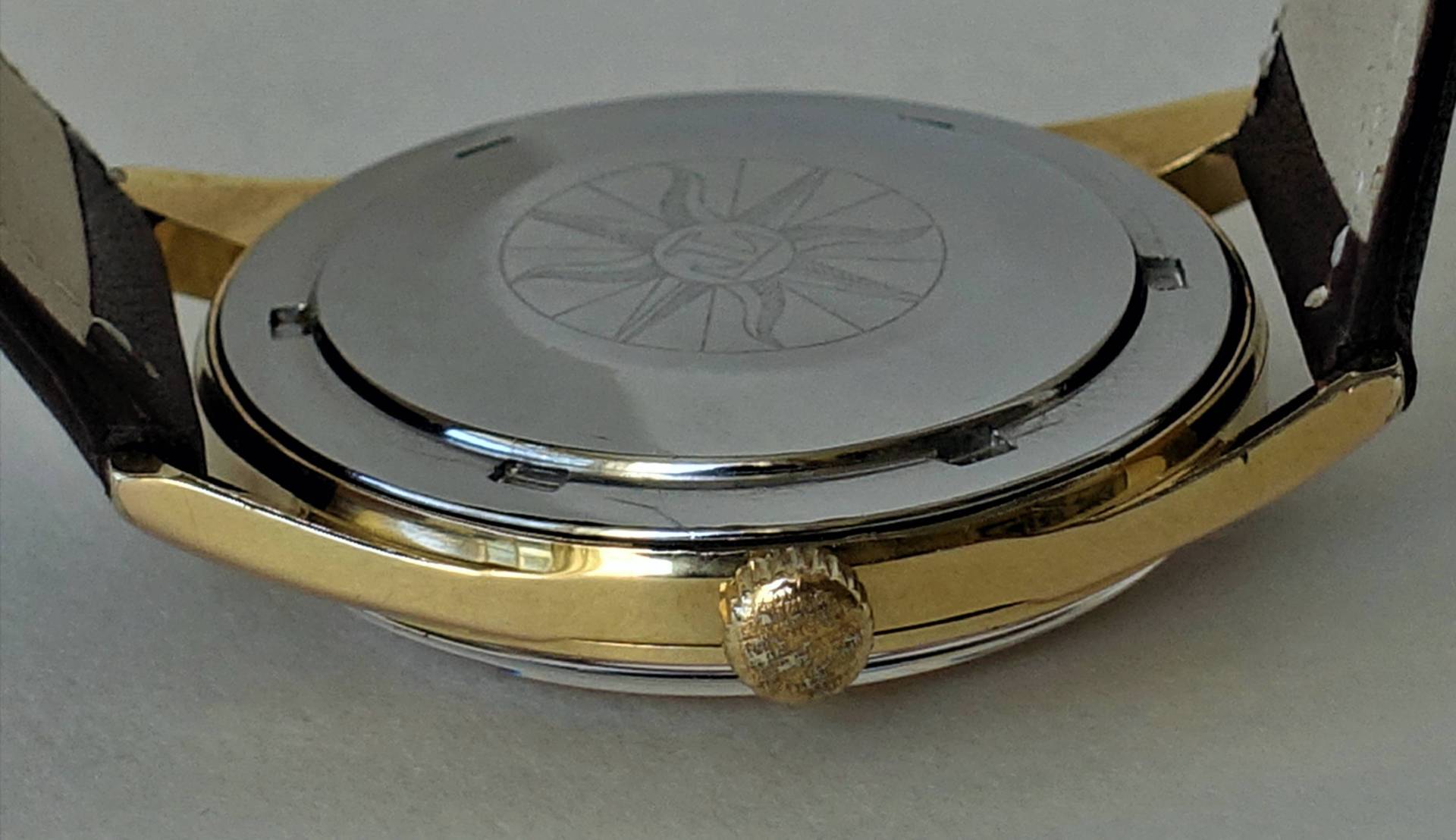 Rückseite einer Armbanduhr mit silberfarbenem Boden und goldfarbener Gehäusekante, seitlich eine goldfarbene Krone.