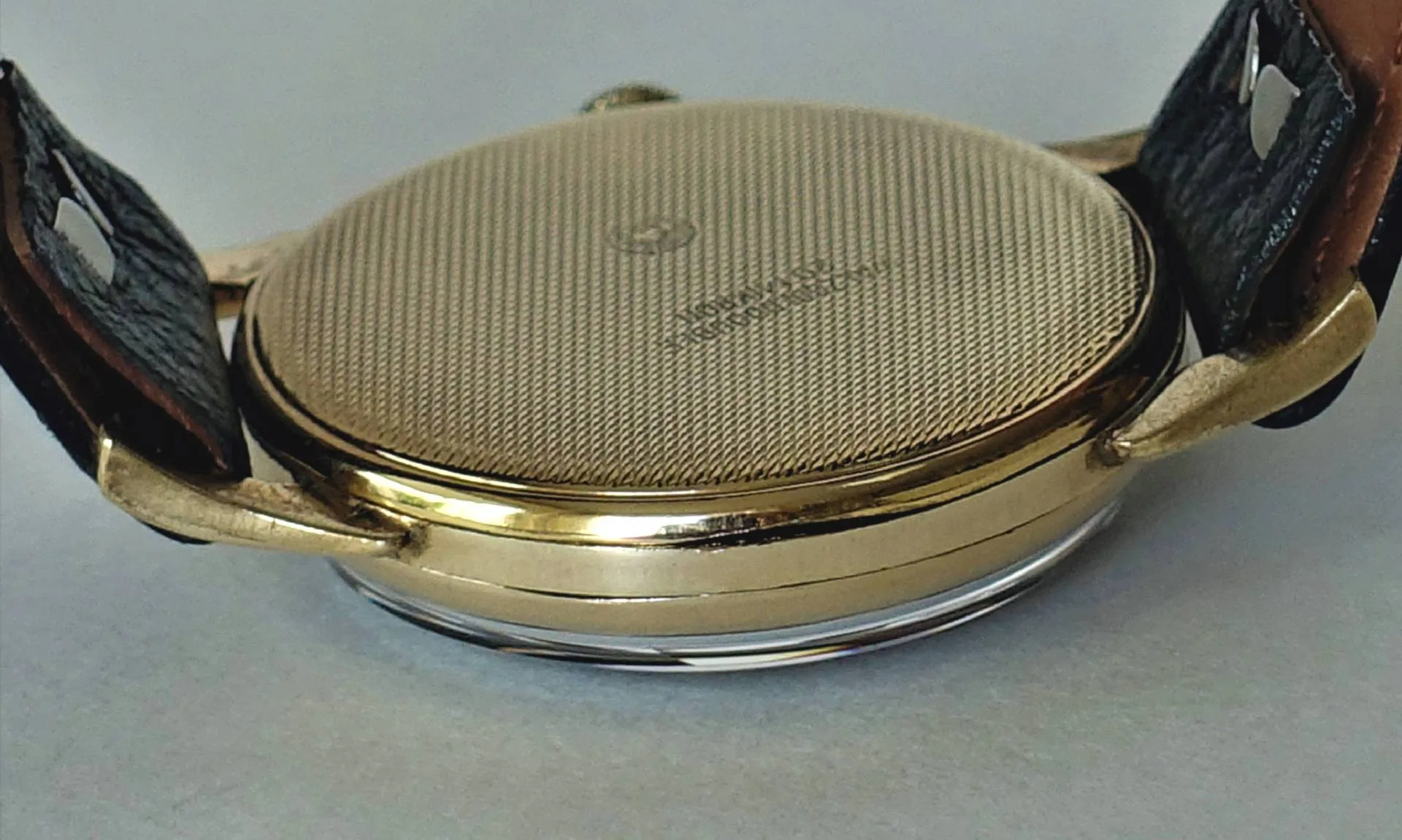 Gelb glänzendes Uhrengehäuse Rückseite Die Rückseite einer Armbanduhr mit fein guillochiertem, gelbfarben glänzendem Gehäuse und schwarzem Lederarmband ist zu sehen.