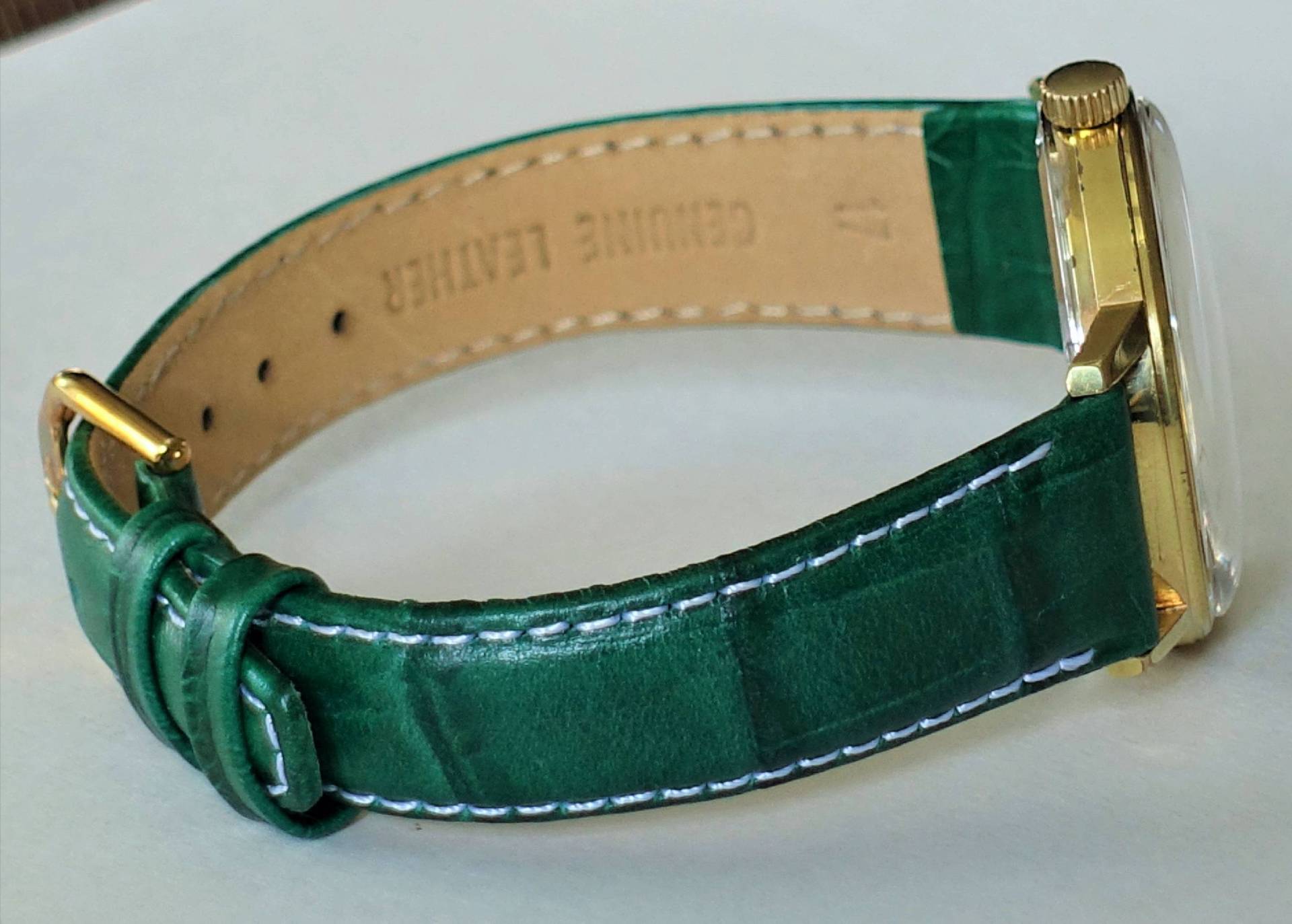 Eine Armbanduhr mit grünem Lederarmband, weißer Naht und goldfarbenem Gehäuse liegt auf einer hellen Oberfläche.