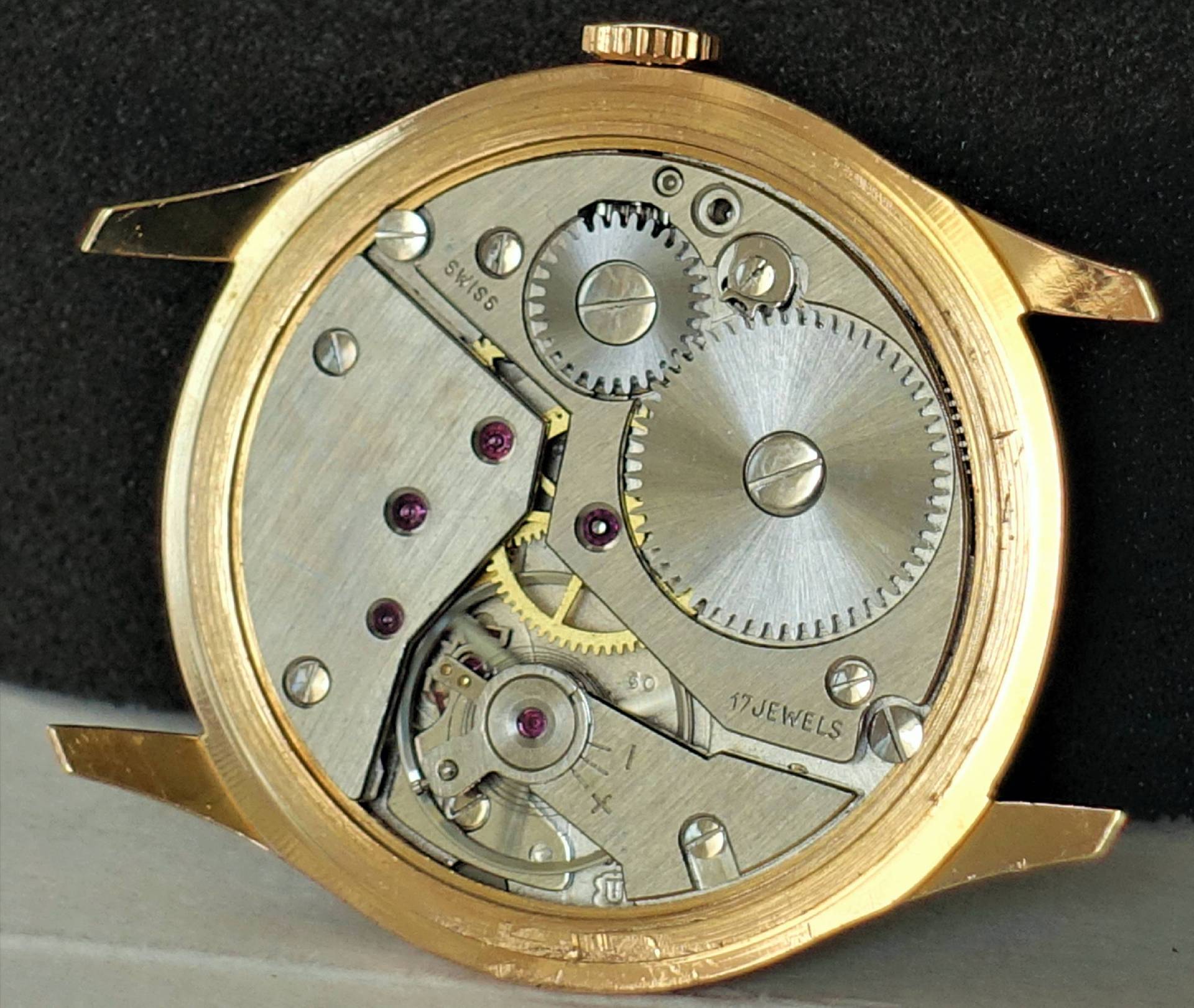 Geöffnetes Uhrgehäuse in Goldfarbe zeigt ein mechanisches Uhrwerk mit silberfarbenen Zahnrädern und sichtbaren roten Lagersteinen.