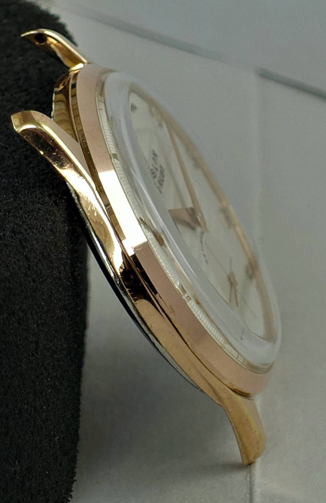 Das Bild zeigt ein seitlich aufgenommenes, poliert roségoldfarbenes Uhrengehäuse mit weißem Zifferblatt und goldfarbenen Zeigern.