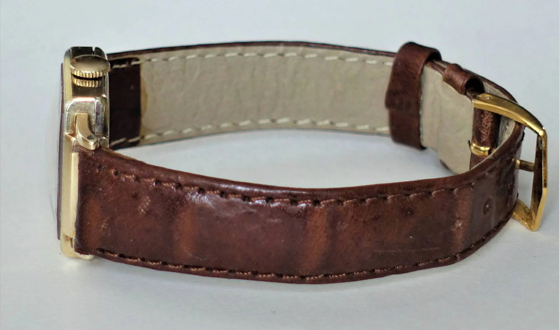 Eine Armbanduhr mit braunem Lederarmband und gelb-metallenem Gehäuse sowie Schließe, von der Seite fotografiert.