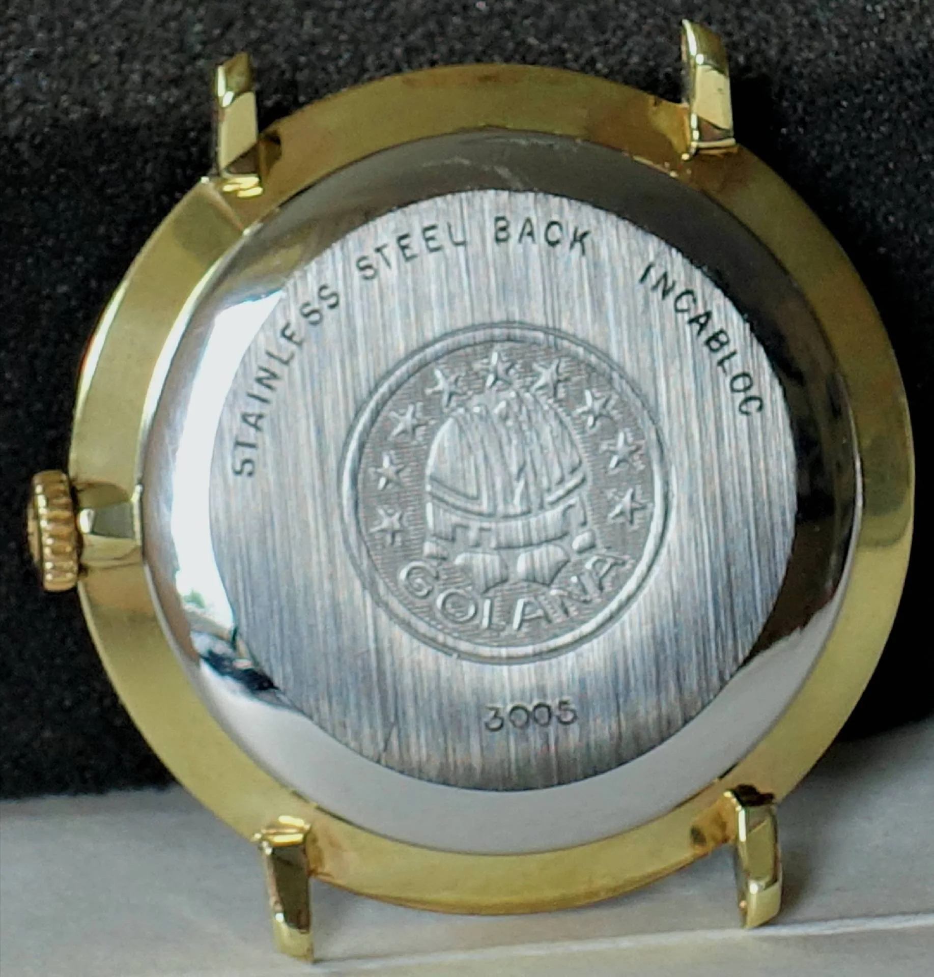 Rundes Uhrengehäuse mit silberfarbener Rückseite und goldfarbener Lünette, Gravur und Krone an der Seite.