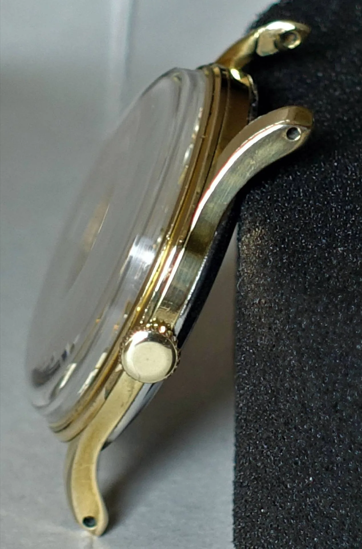 Seitlich aufgenommenes goldfarbenes Uhrengehäuse mit Krone und Glas.