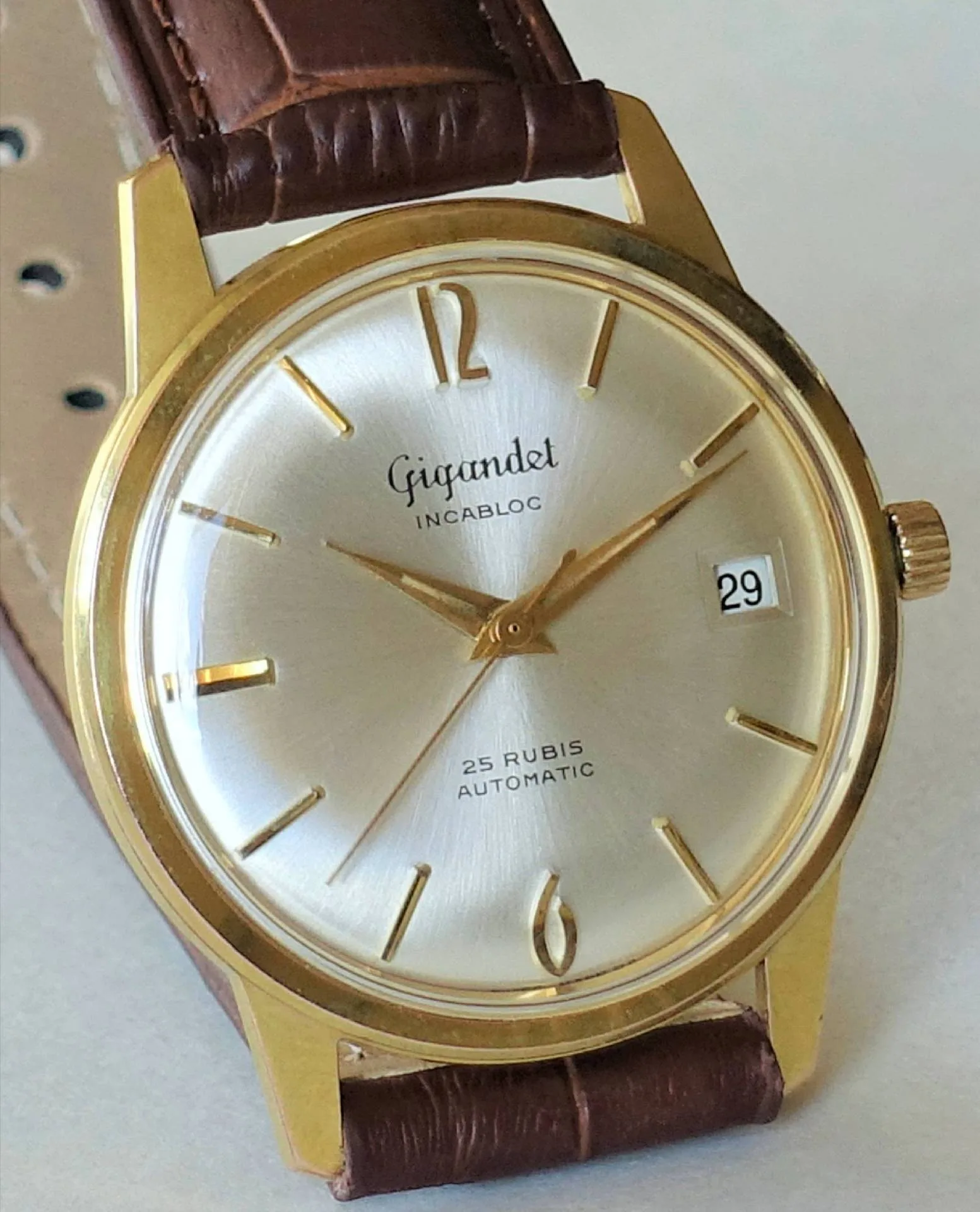 Goldfarben Silber Zifferblatt Braunband Eine klassische Armbanduhr mit goldfarbenem Gehäuse, silbernem Zifferblatt, braunem Lederarmband und Datumsanzeige bei drei Uhr.
