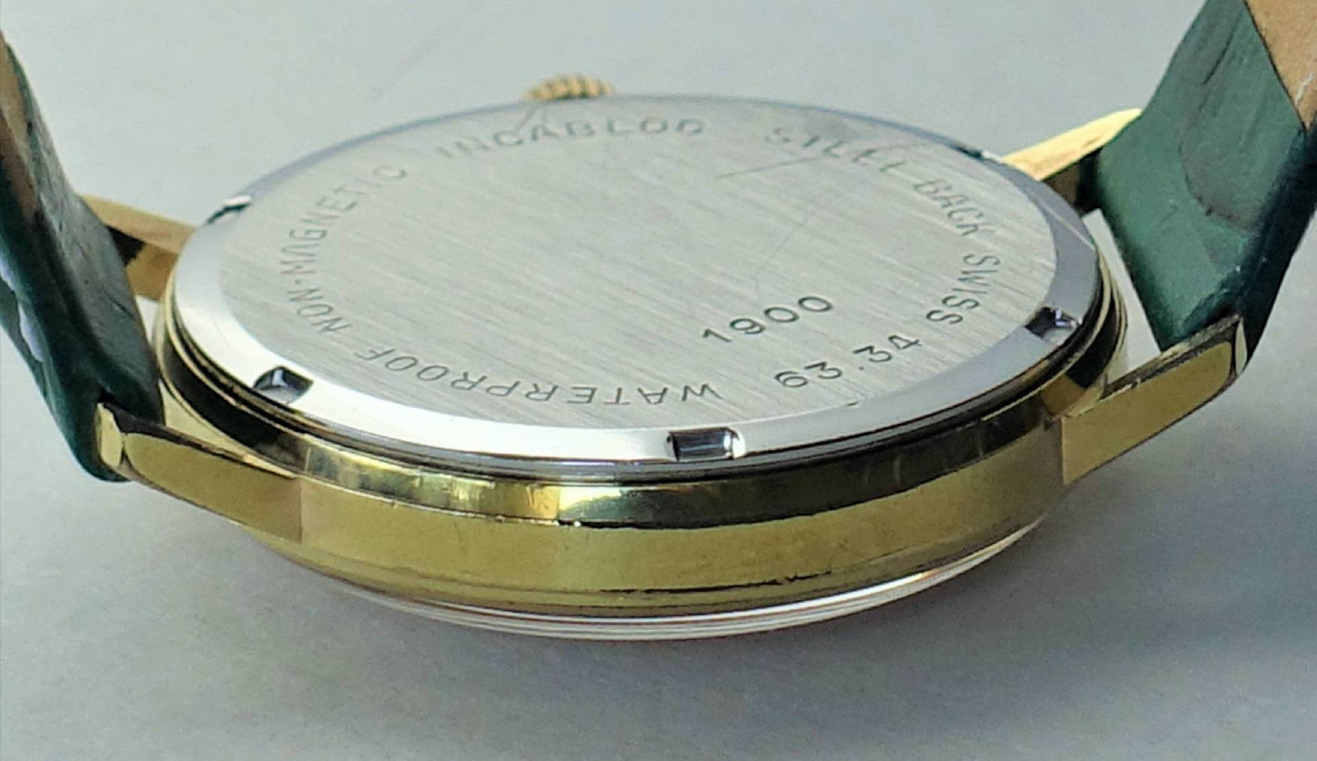 Silberfarbener runder Uhrenboden mit Gravur, montiert an einem goldfarbenen Uhrengehäuse mit grünem Armband.