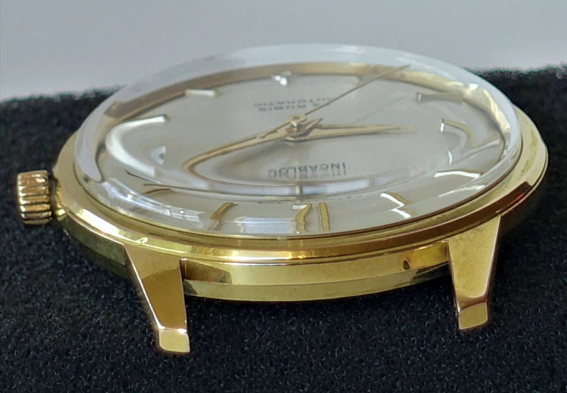 Eine Armbanduhr mit glänzend gelbgoldenem Gehäuse und gewölbtem Glas liegt auf schwarzem Stoff, das Zifferblatt ist silbern mit goldfarbenen Zeigern und Indizes.