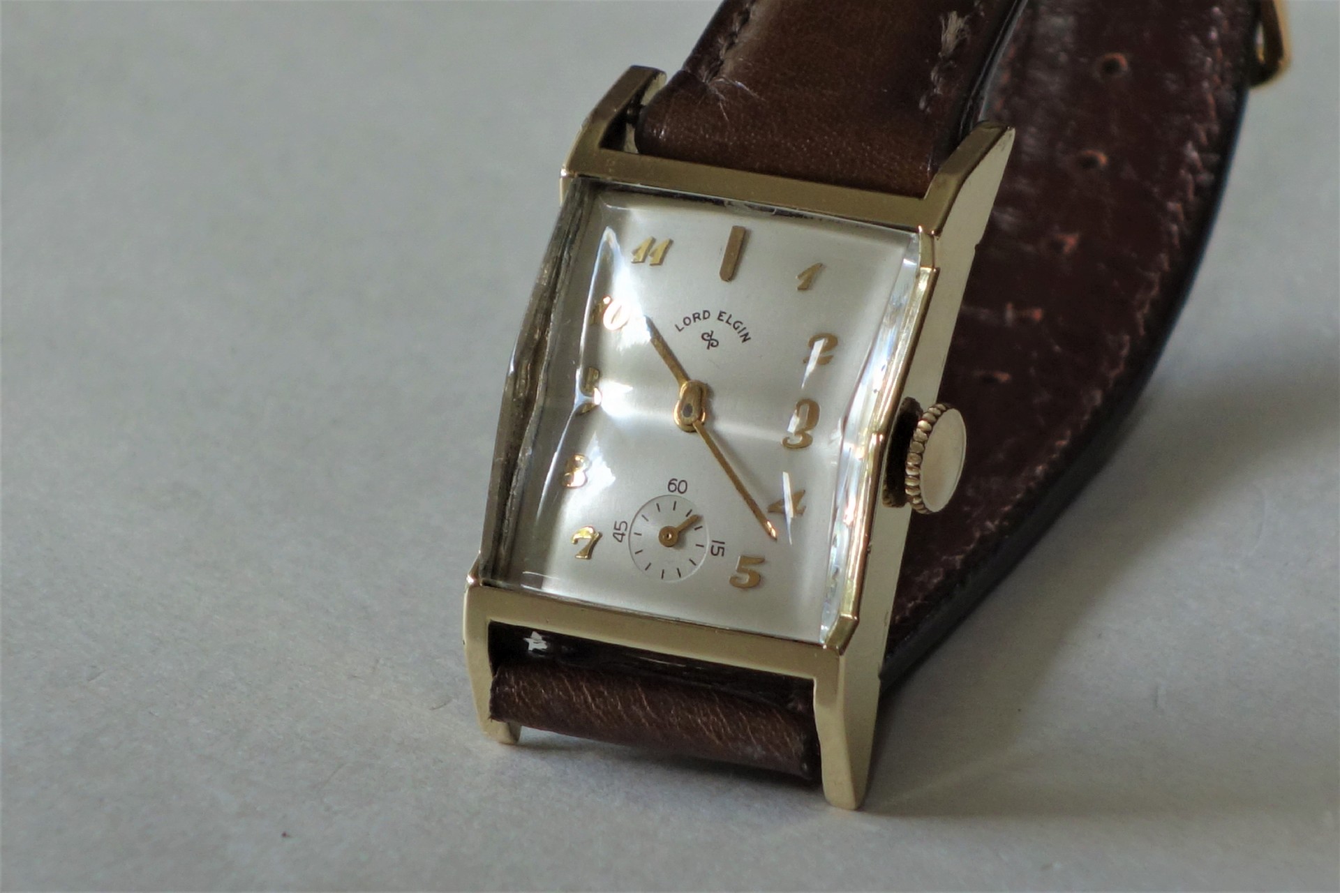 Rechteckige Armbanduhr mit braunem Armband und hellem Zifferblatt.