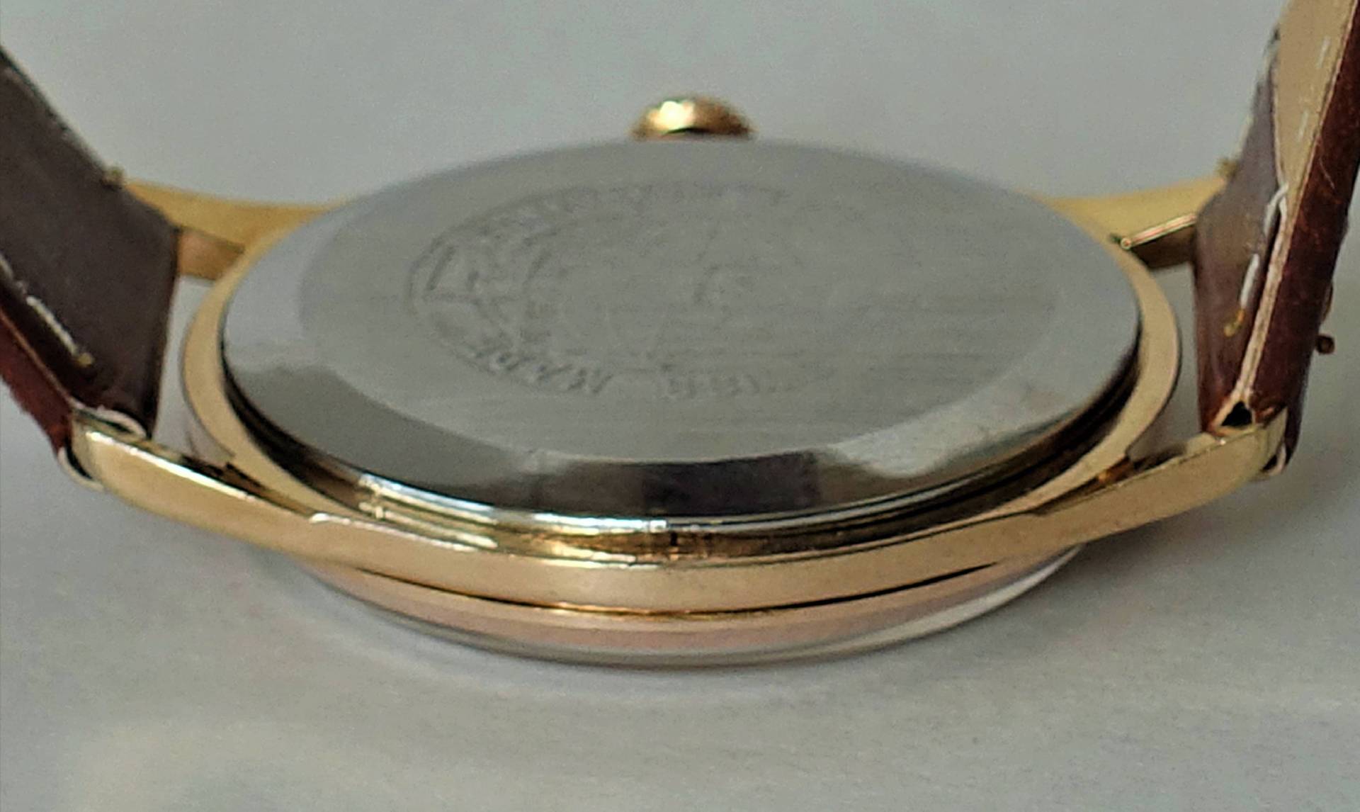 Das Bild zeigt die Rückseite einer Armbanduhr mit einem silberfarbenen Gehäuseboden und einem goldfarbenen Gehäuserand sowie braunem Lederarmband.