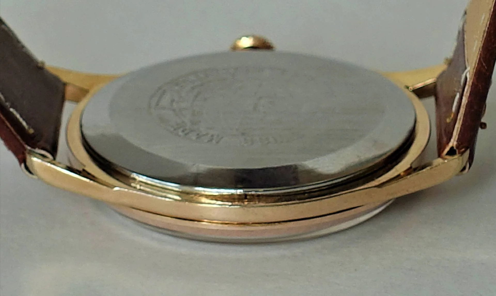Das Bild zeigt die Rückseite einer Armbanduhr mit einem silberfarbenen Gehäuseboden und einem goldfarbenen Gehäuserand sowie braunem Lederarmband.