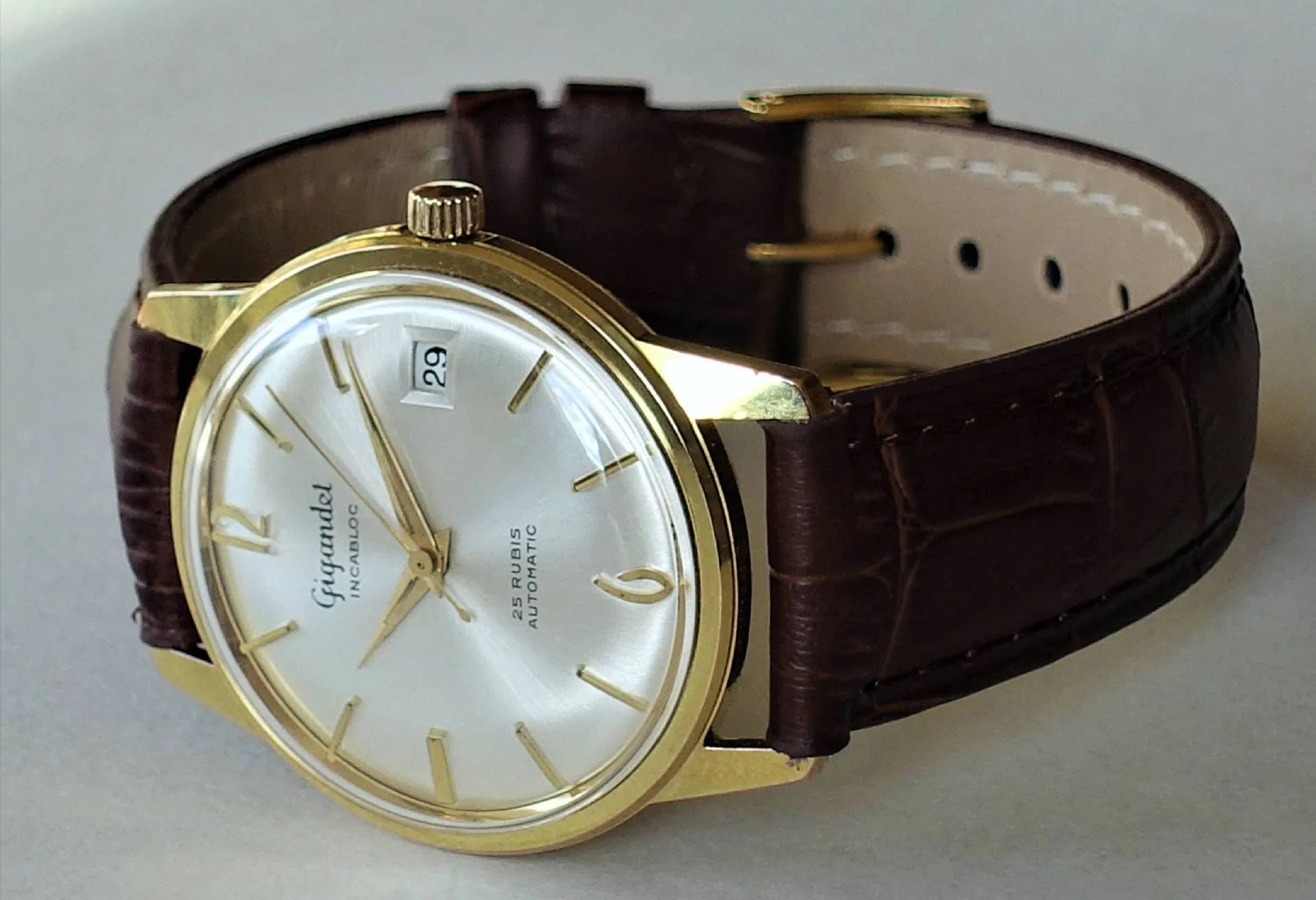 Goldfarbenes Gehäuse, braunes Lederband Eine Armbanduhr mit goldfarbenem Gehäuse, silberfarbenem Zifferblatt und braunem Lederarmband liegt auf einer hellen Oberfläche.