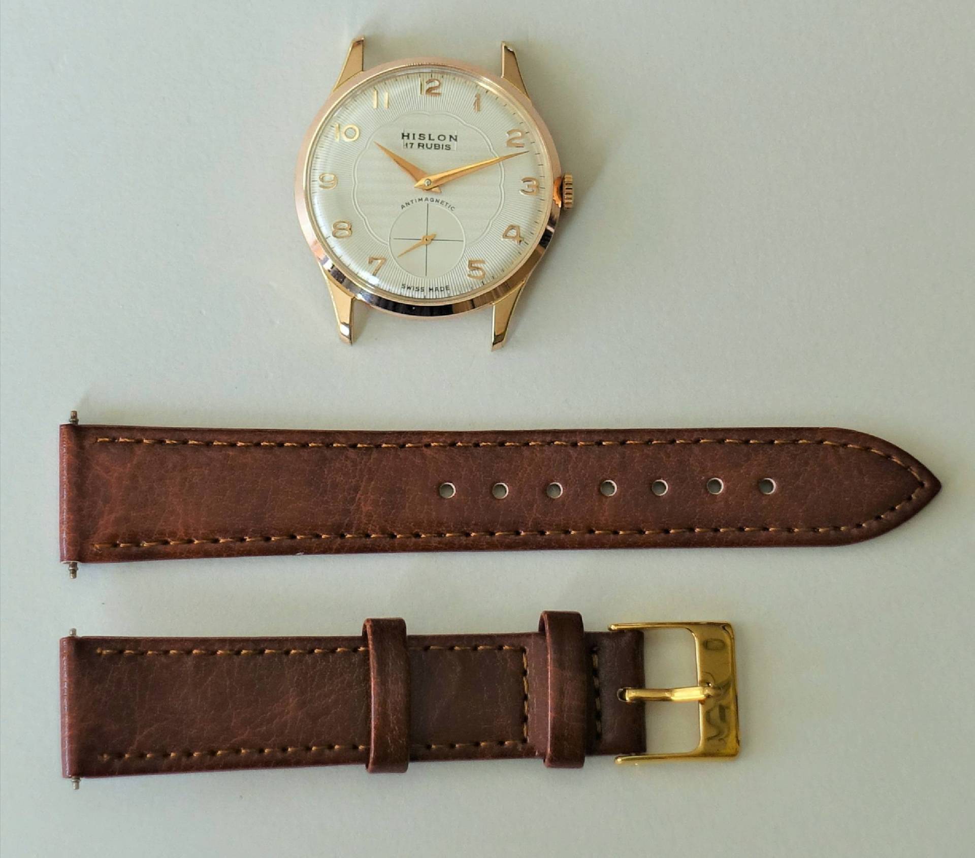 Eine Armbanduhr mit weiß-goldenem Gehäuse und separatem braunen Lederarmband mit goldfarbener Schließe liegt auf hellem Untergrund.