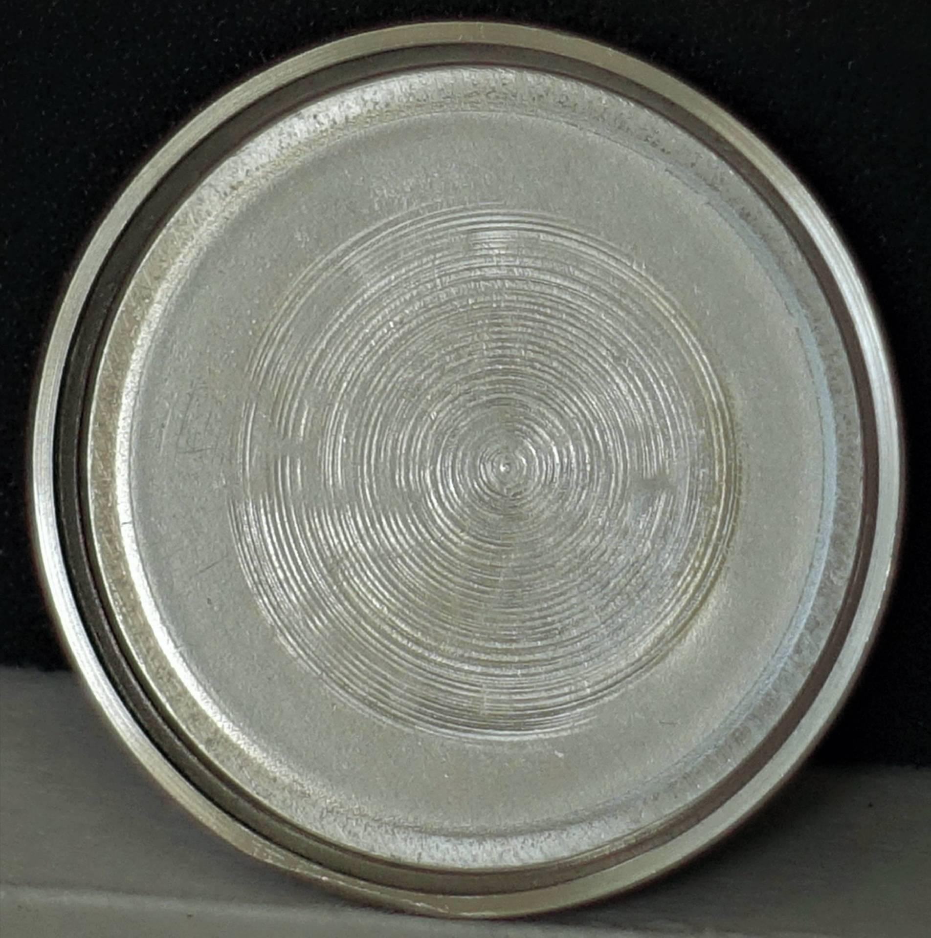 Rundes, silberfarbenes Uhrendeckel-Innenteil mit konzentrischem Kreismuster auf der Oberfläche.