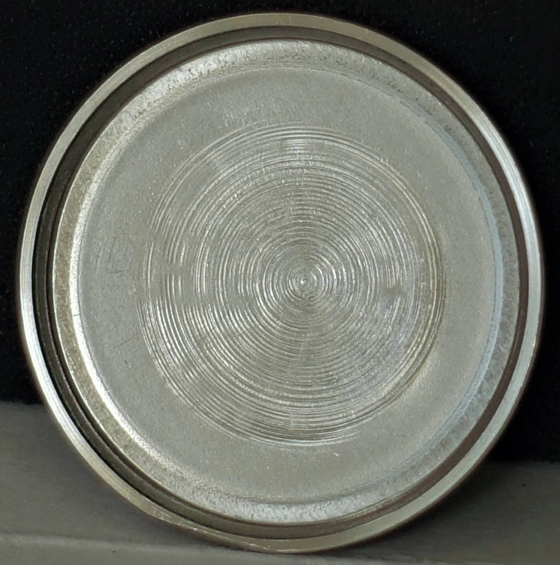 Rundes, silberfarbenes Uhrendeckel-Innenteil mit konzentrischem Kreismuster auf der Oberfläche.