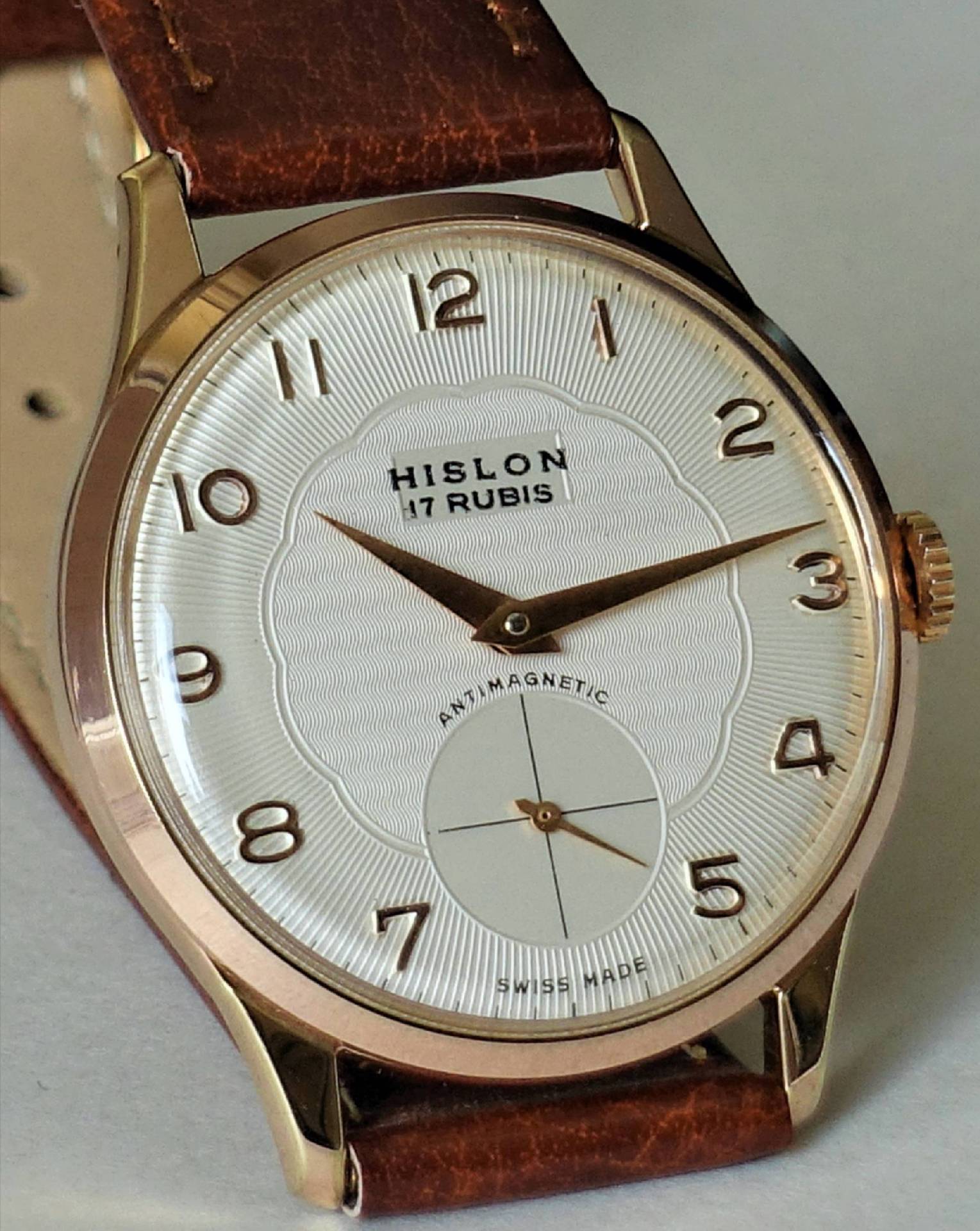 Eine klassische Armbanduhr mit weißem Zifferblatt, goldfarbenem Gehäuse und braunem Lederarmband, auf dem Zifferblatt steht "HISLON 17 RUBIS".