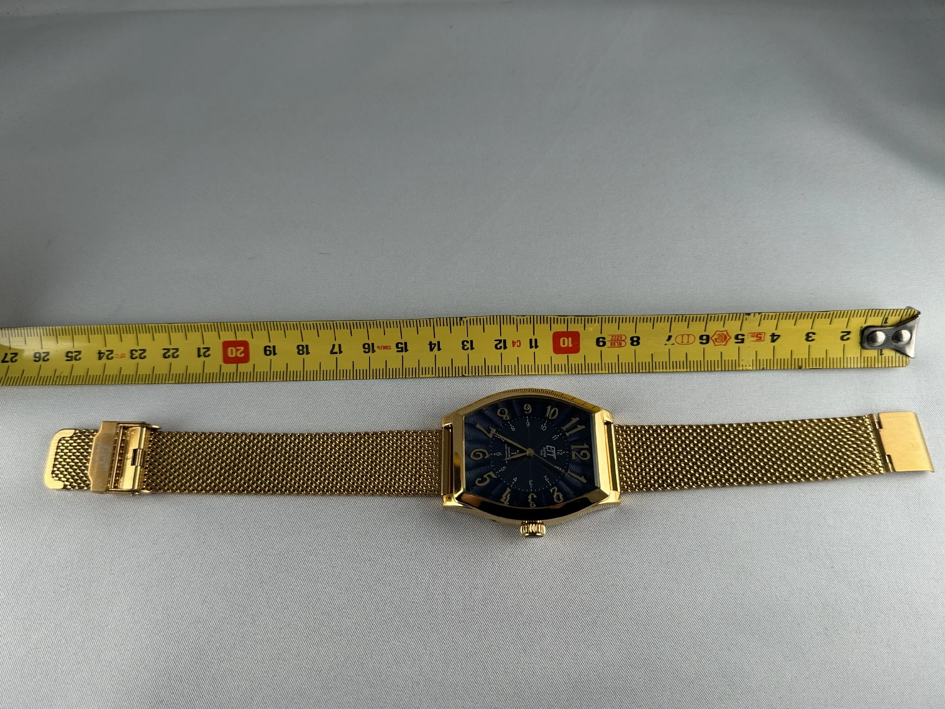 Goldfarbene Armbanduhr mit schwarzem Zifferblatt und Milanaise-Edelstahlarmband, daneben ein Maßband.