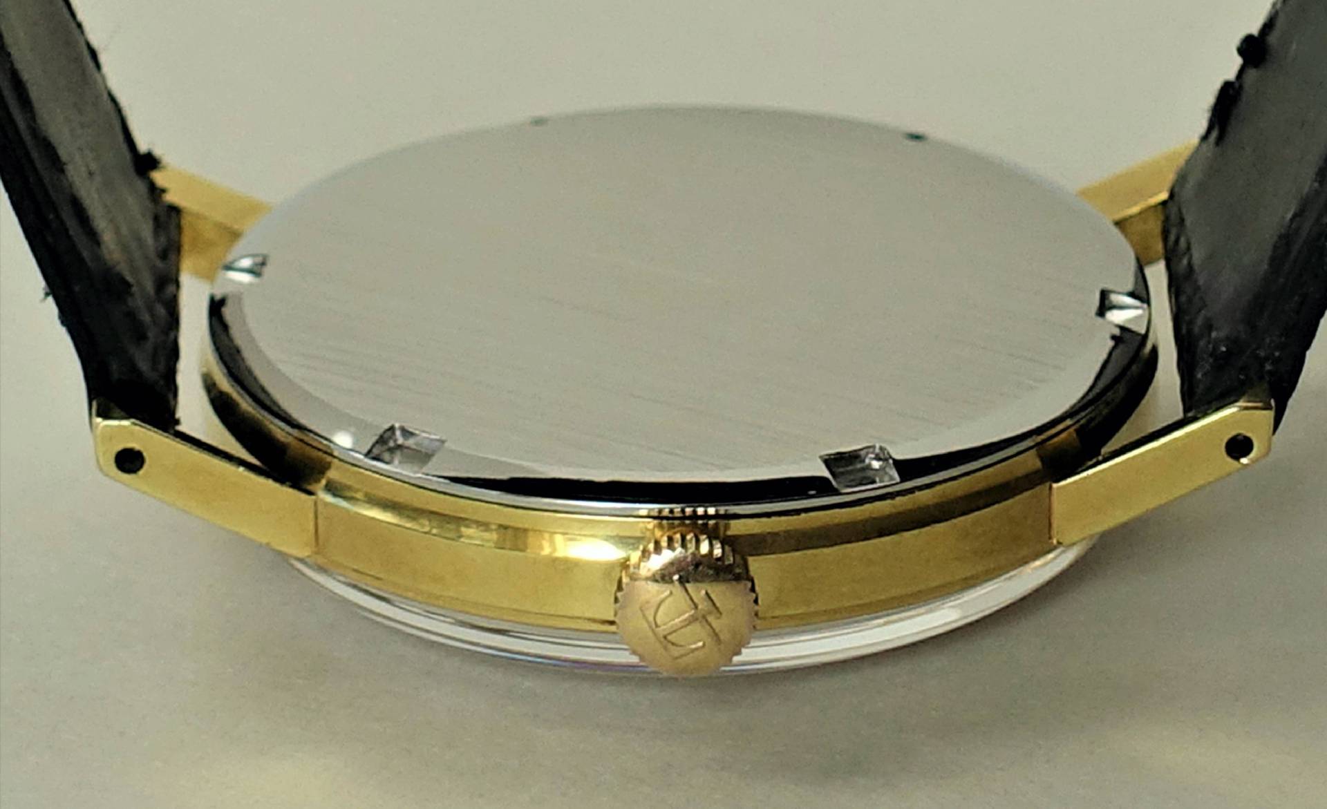 Rundes Uhrengehäuse in goldfarbener Optik mit silberfarbener Rückseite und seitlicher Krone.