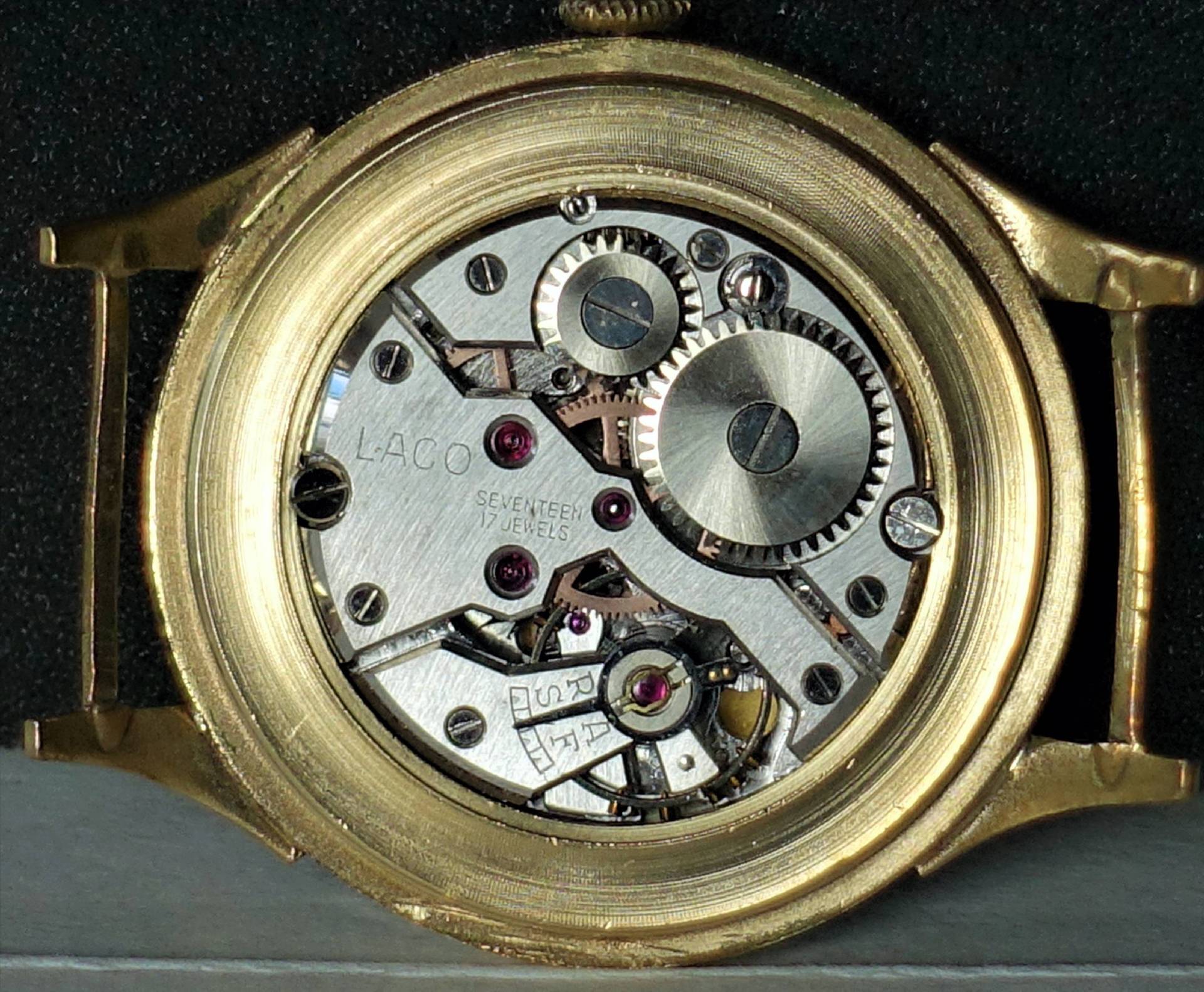 Silberfarbenes mechanisches Uhrwerk mit sichtbaren Zahnrädern in einem goldfarbenen Gehäuse.