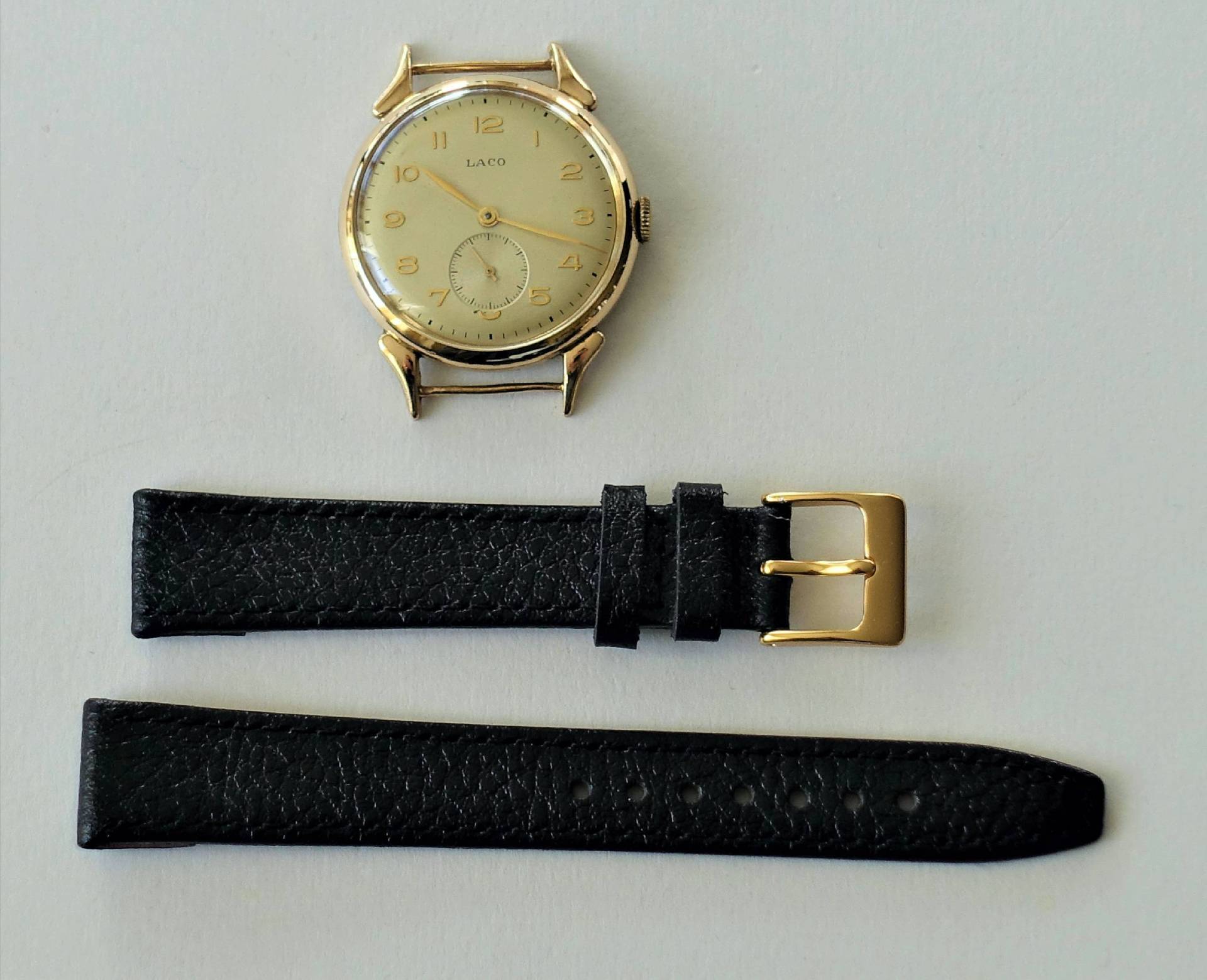 Goldfarbene Armbanduhr mit hellem Zifferblatt und schwarzem Lederarmband mit goldfarbener Schließe.