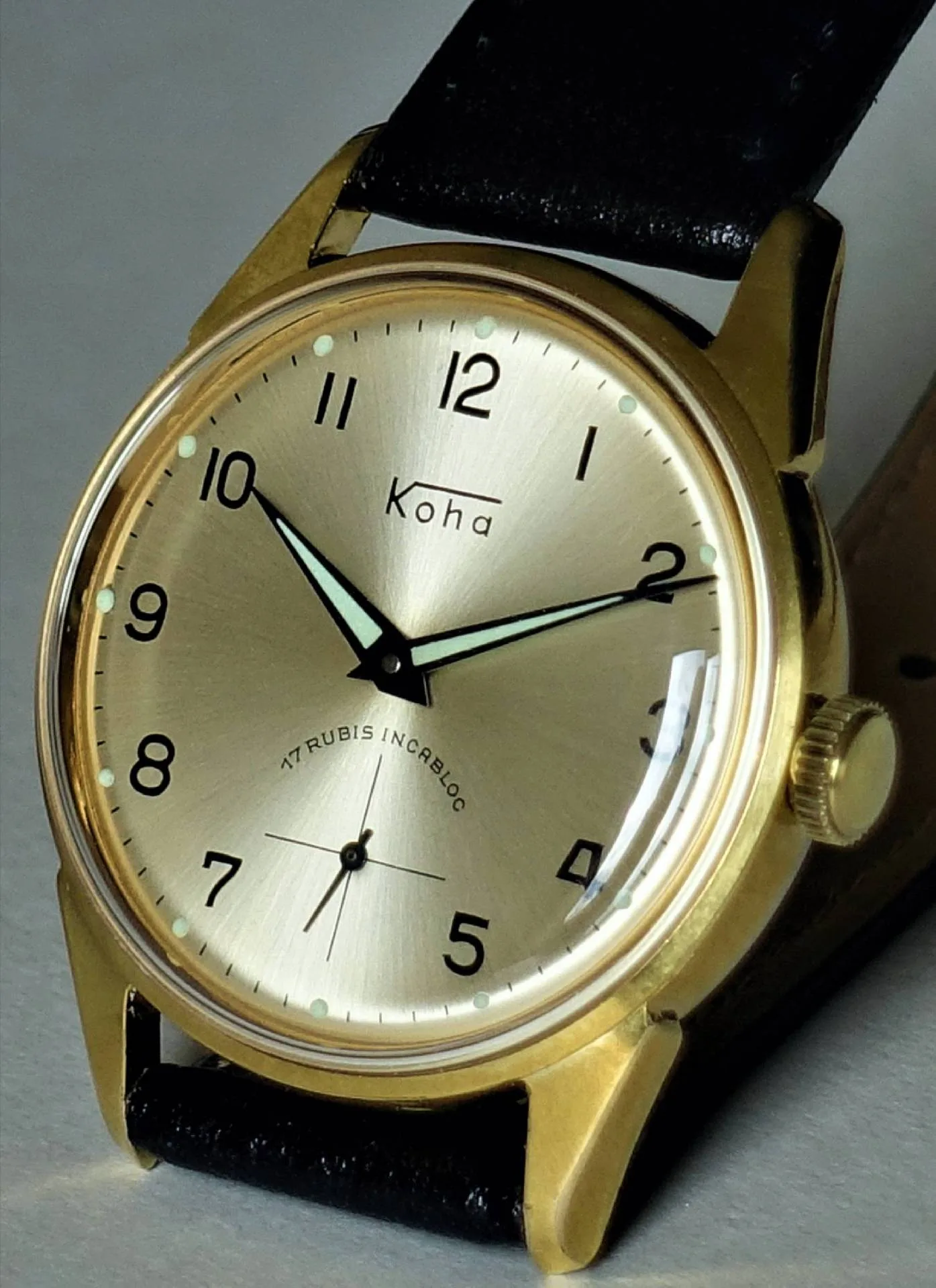 Goldfarbene Armbanduhr mit hellem Zifferblatt, schwarzen Zahlen und schwarzem Lederarmband.