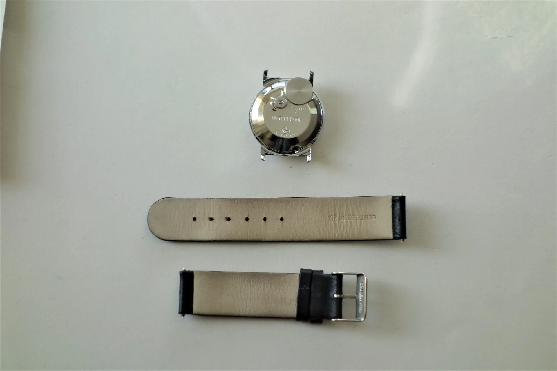 Zerlegtes Uhrgehäuse aus Metall mit zwei schwarzen Lederarmbandteilen und Metallschließe.