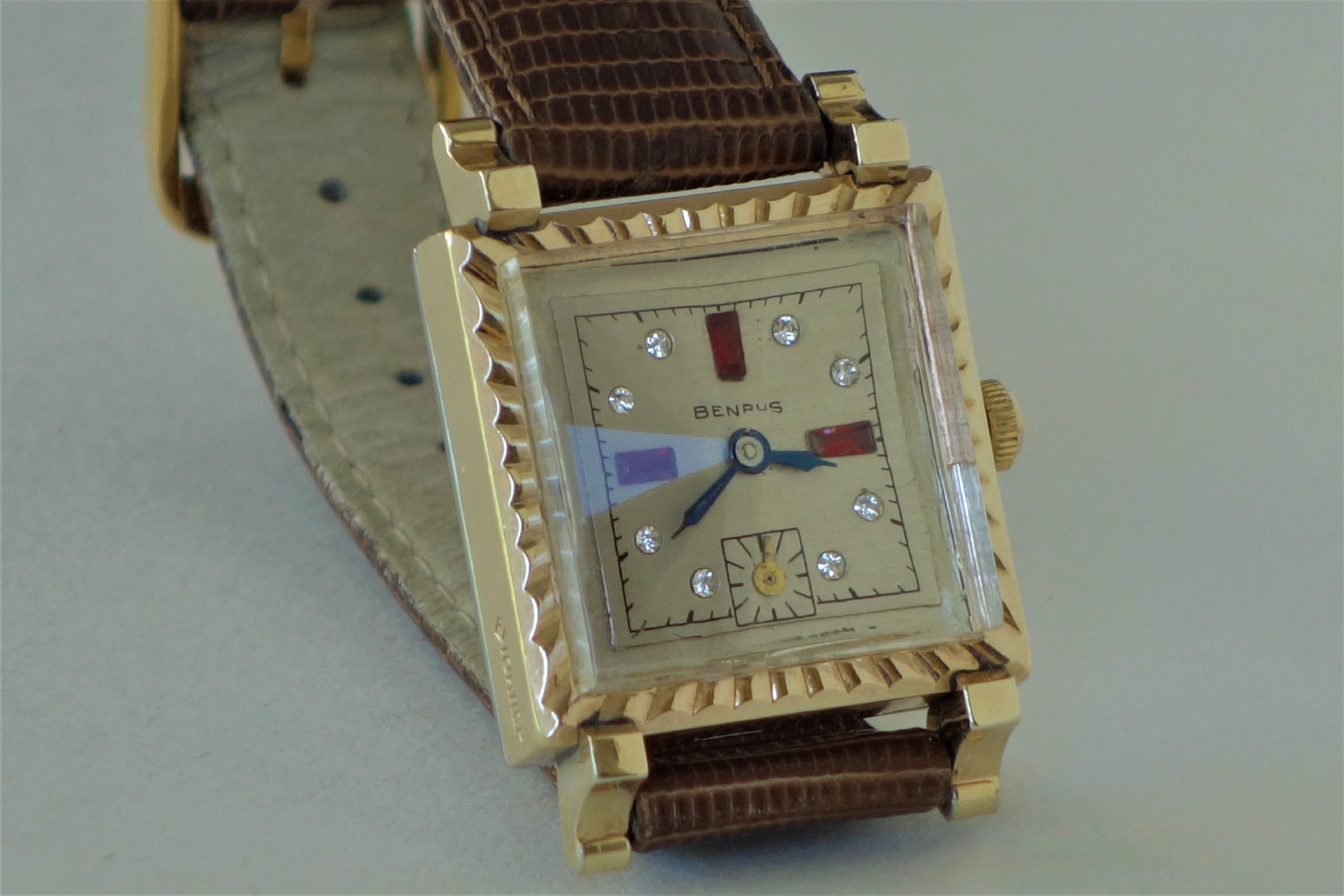 Rechteckige Armbanduhr mit goldfarbenem Gehäuse, braunem Lederarmband und verziertem Zifferblatt.
